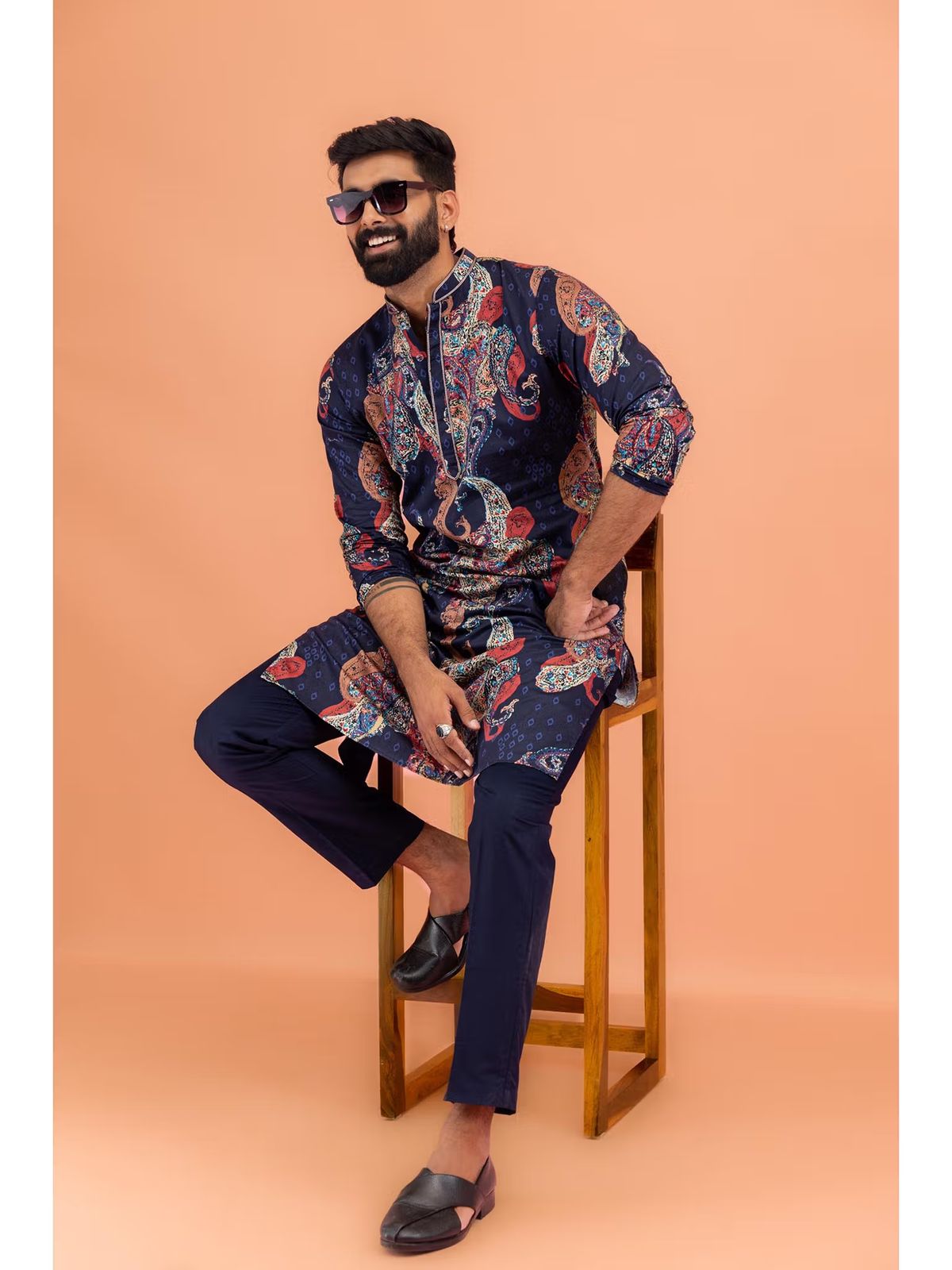 Male model seated on wooden stool wearing men's black printed kurta Canada with navy pants and loafers