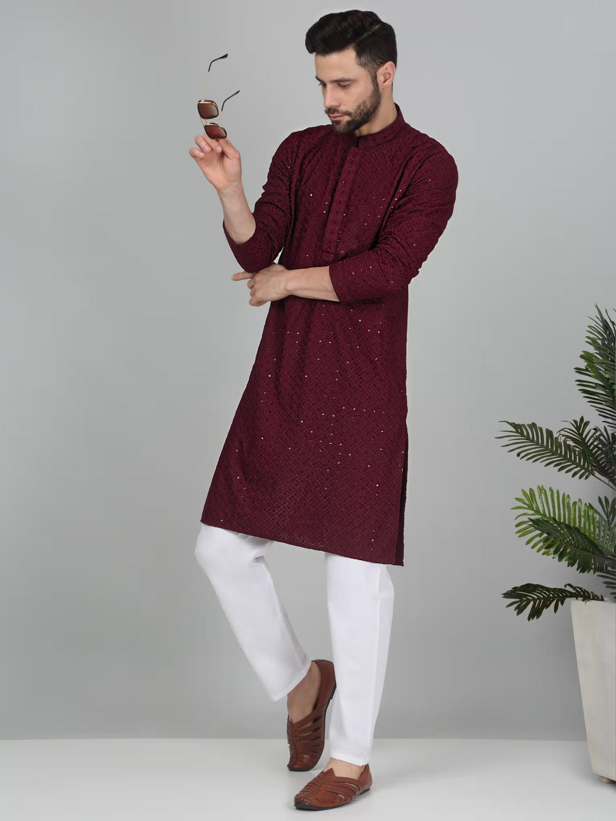 Male model wearing men's burgundy embroidered kurta set with white trousers and brown loafers holding sunglasses