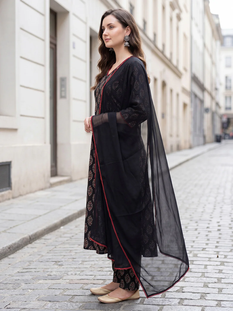 A woman stands on a cobblestone street wearing a black embroidered kurta set with a matching sheer dupatta. The outfit features intricate gold and red floral embroidery, and she is wearing beige flat shoes.