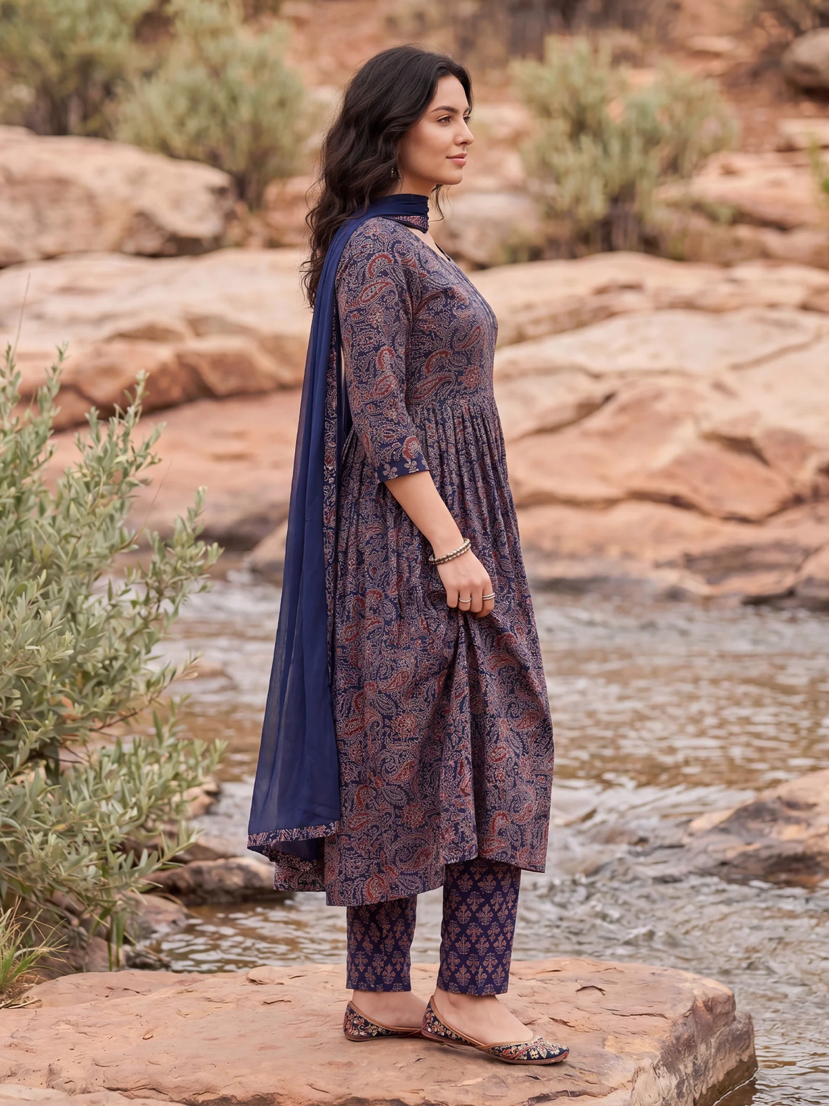 A woman stands outdoors by a rocky stream wearing a dark patterned kurta with a navy dupatta and matching patterned pants. The setting includes reddish-brown rocks and green shrubs.