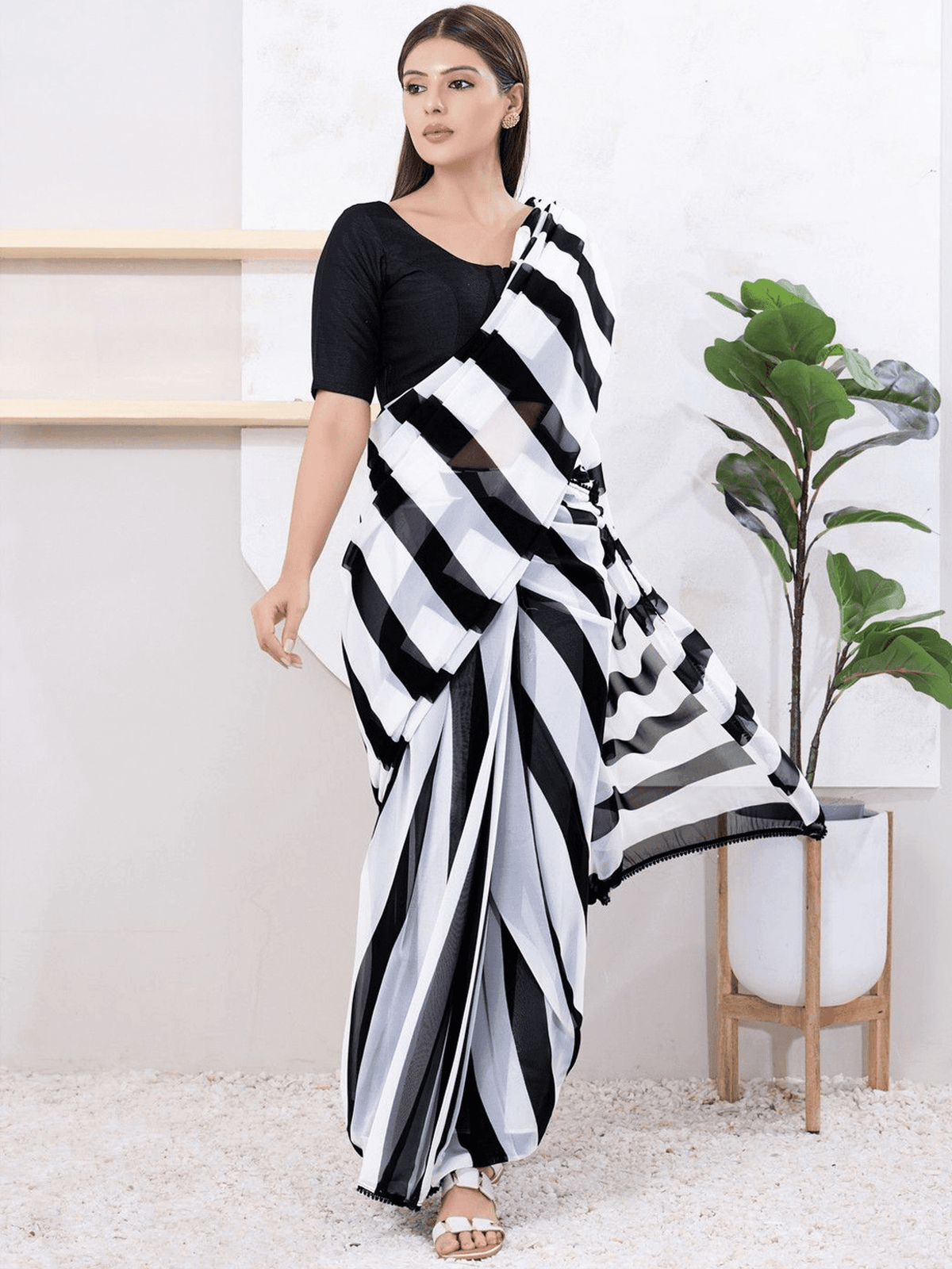 A woman modeling a black and white striped sari with a solid black short-sleeve blouse, standing in a bright indoor setting with a potted plant.