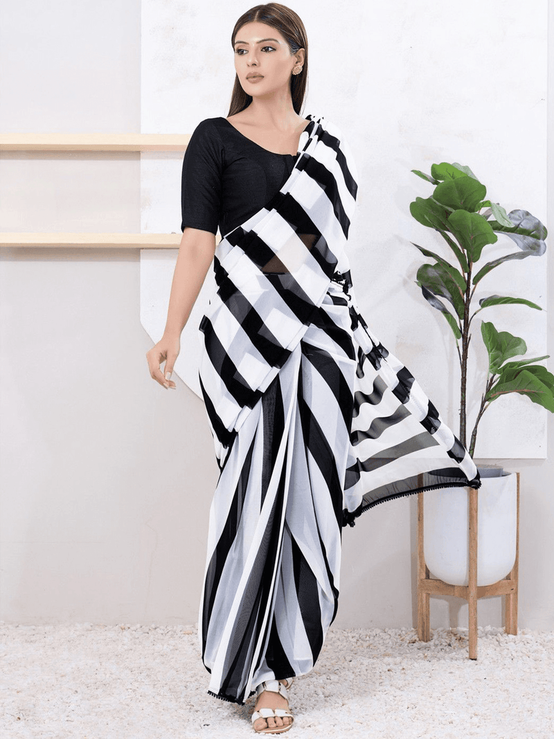 A woman modeling a black and white striped sari with a solid black short-sleeve blouse, standing in a bright indoor setting with a potted plant.