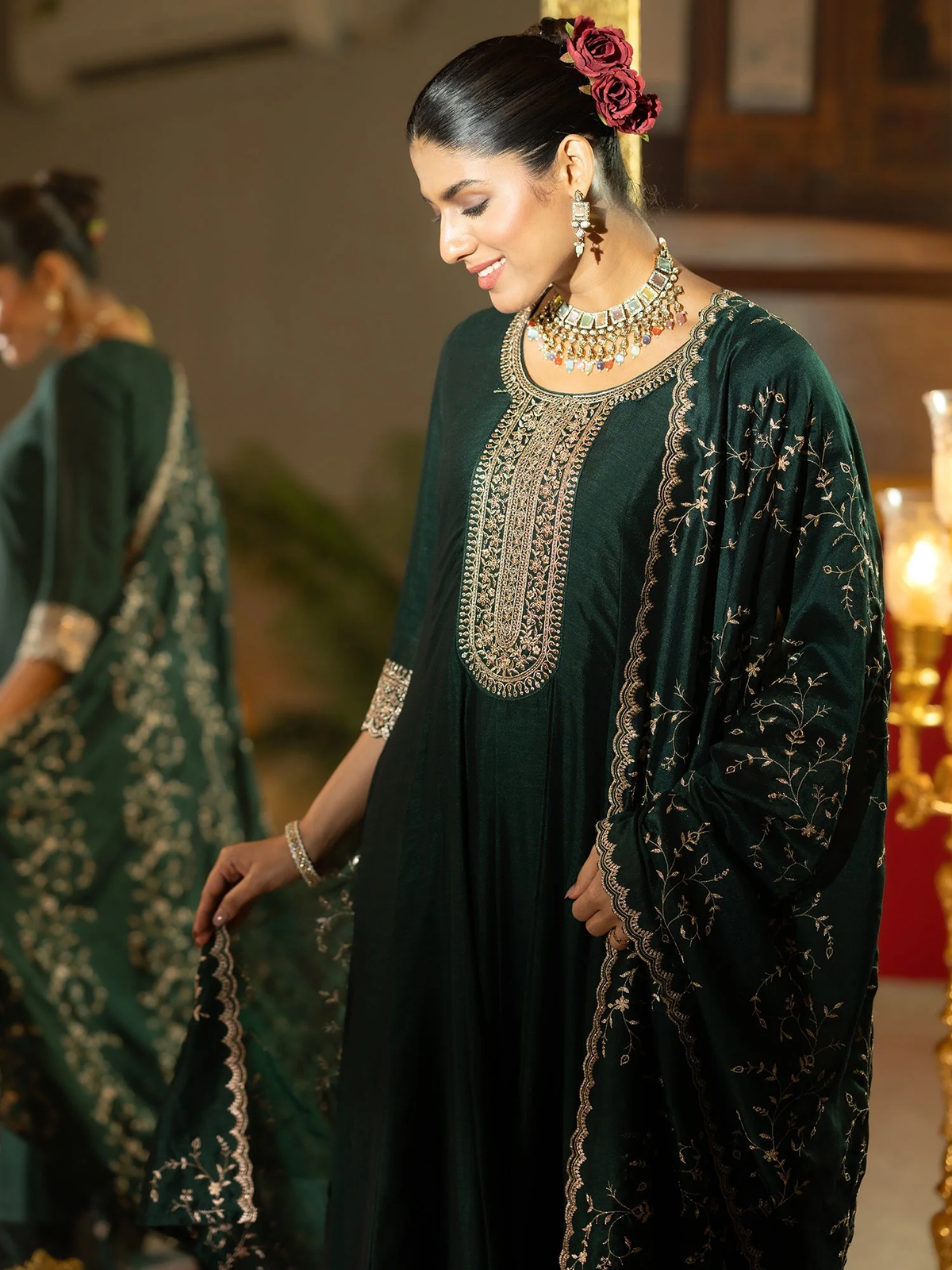 Woman in a dark green embroidered traditional outfit with jewelry, standing indoors.