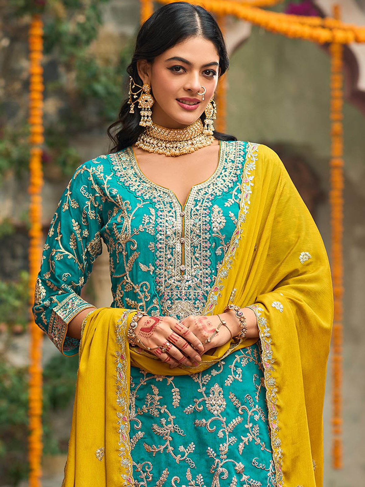 Woman in traditional turquoise and yellow embroidered outfit with jewelry, standing against a blurred background.