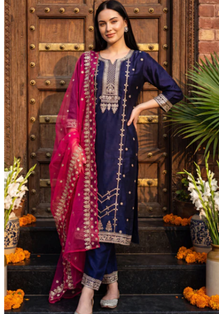A woman models a navy blue and magenta pink ethnic salwar kameez suit with gold embroidery, standing in front of a rustic wooden door decorated with flowers.
