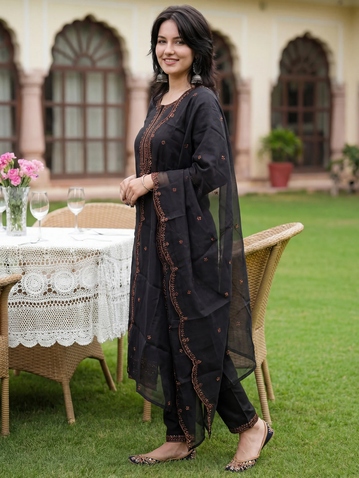 Woman in a black traditional outfit standing outdoors with a table and chairs in the background.