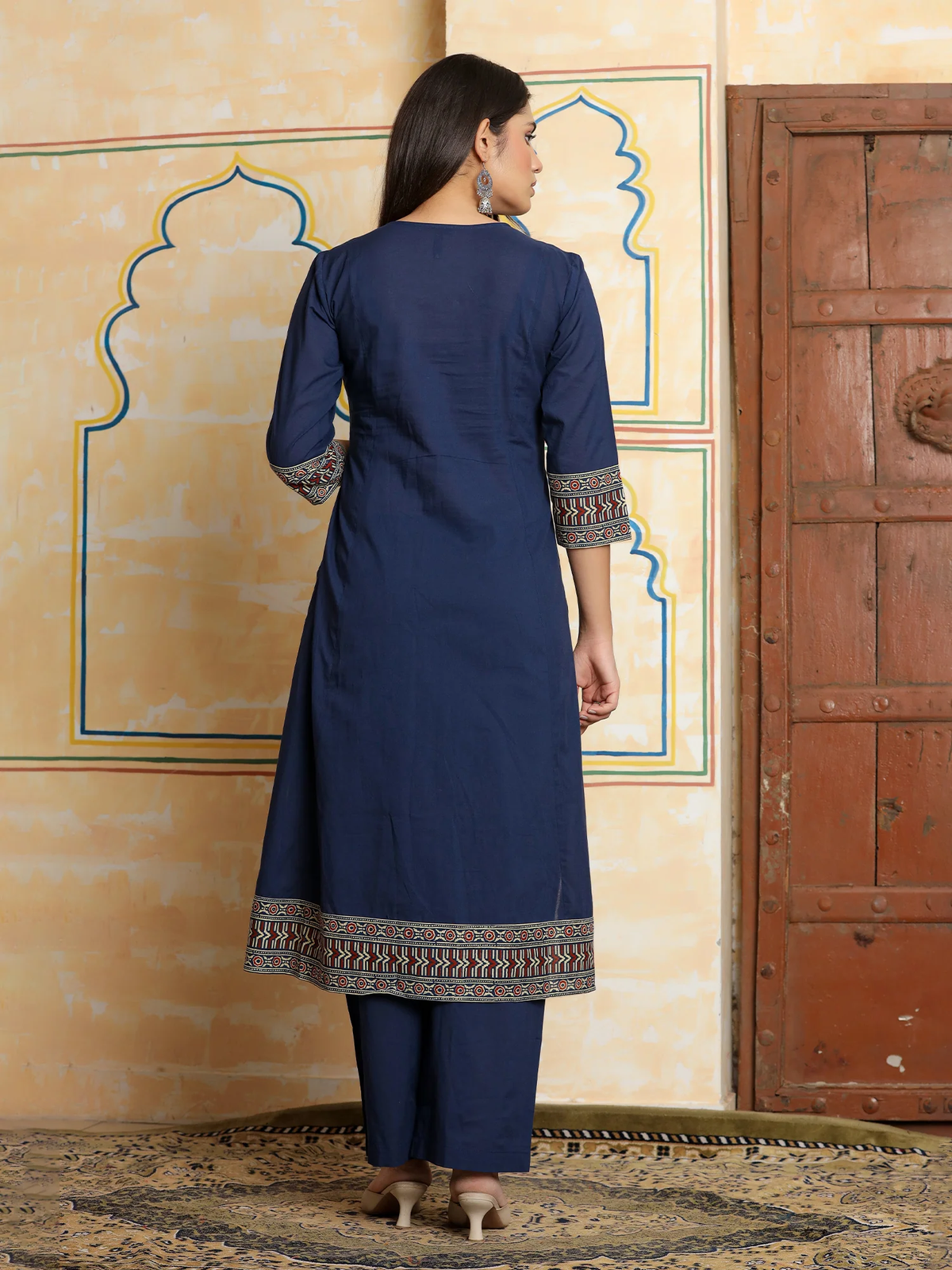 Woman wearing a blue traditional outfit with intricate patterns, standing in front of a decorative wall.