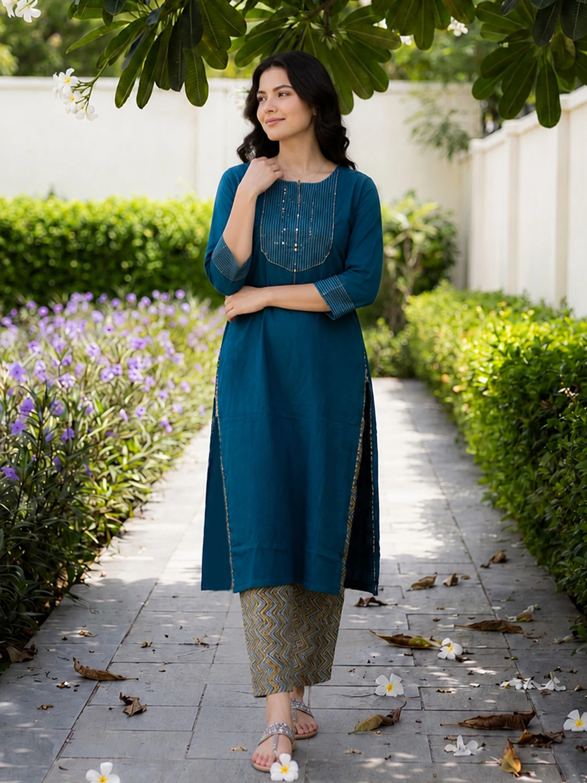 Woman in a teal kurta standing on a pathway with greenery and flowers around.