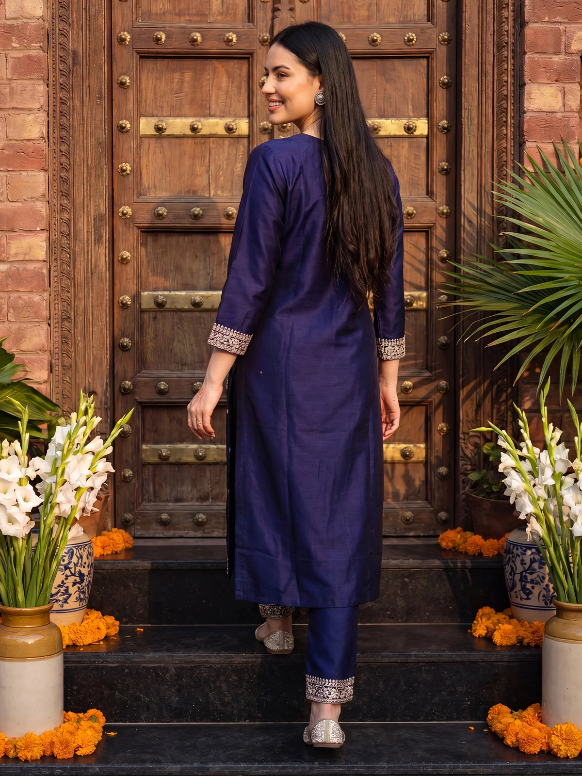 Woman in a blue traditional outfit standing in front of a wooden door with decorative flowers.