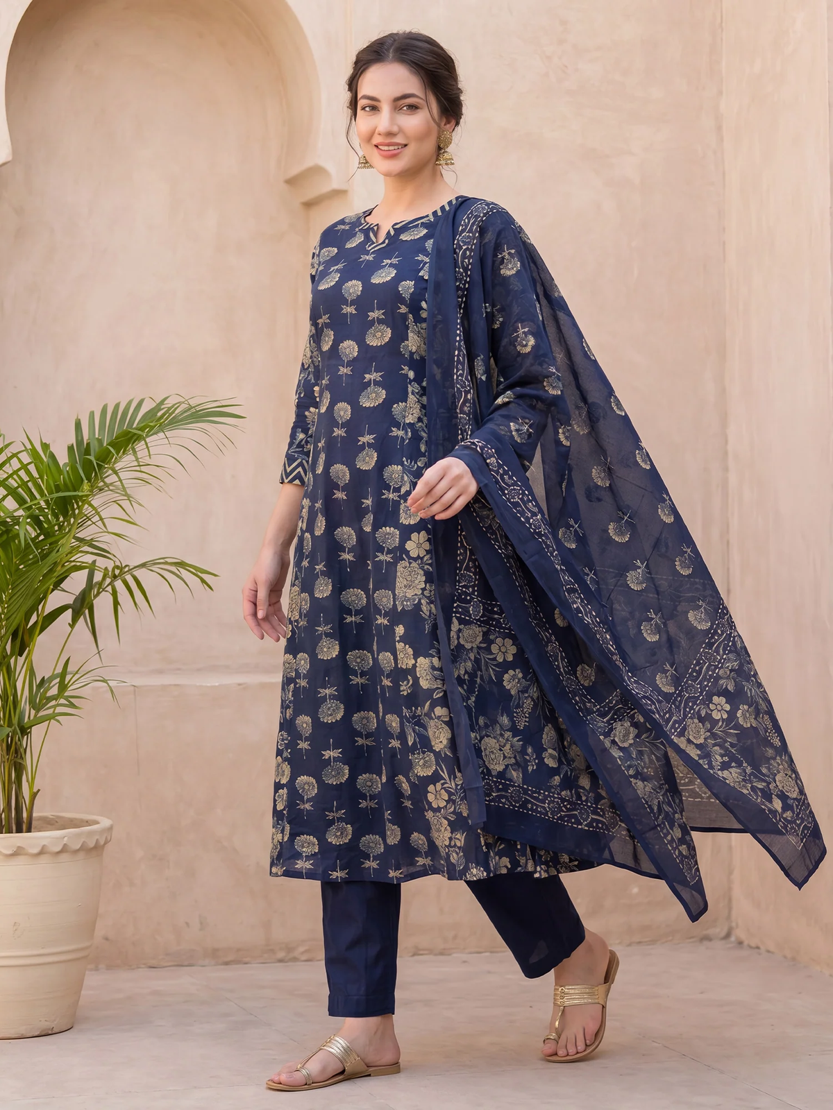 A woman models a navy blue three-piece ethnic suit set featuring a printed kurta, matching trousers, and a sheer printed dupatta, set against a beige stucco wall with a potted plant.