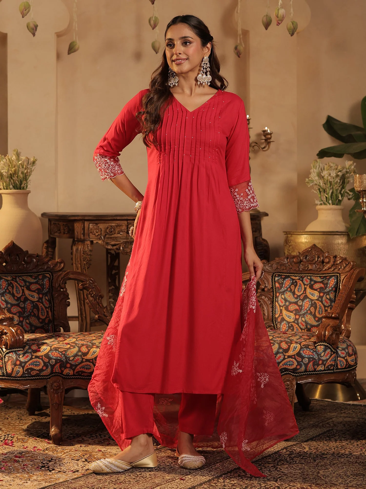 A woman models a bright red three-piece Indian ethnic suit set, featuring a long kurta with pleated detailing and embroidered sleeves, matching trousers, and a sheer embroidered dupatta, set against an ornate traditional interior.