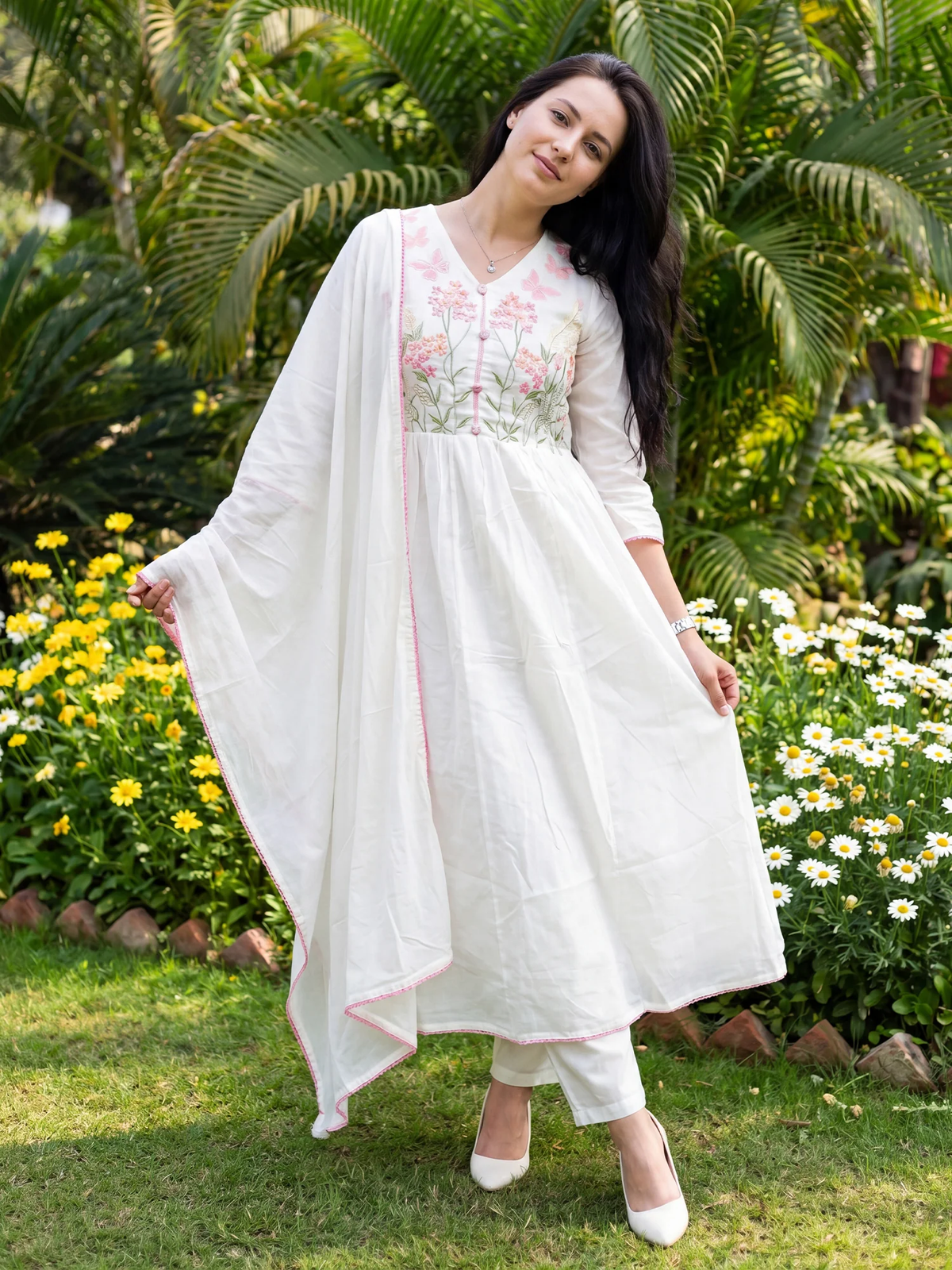 Woman in a white traditional outfit with floral patterns standing in a garden.