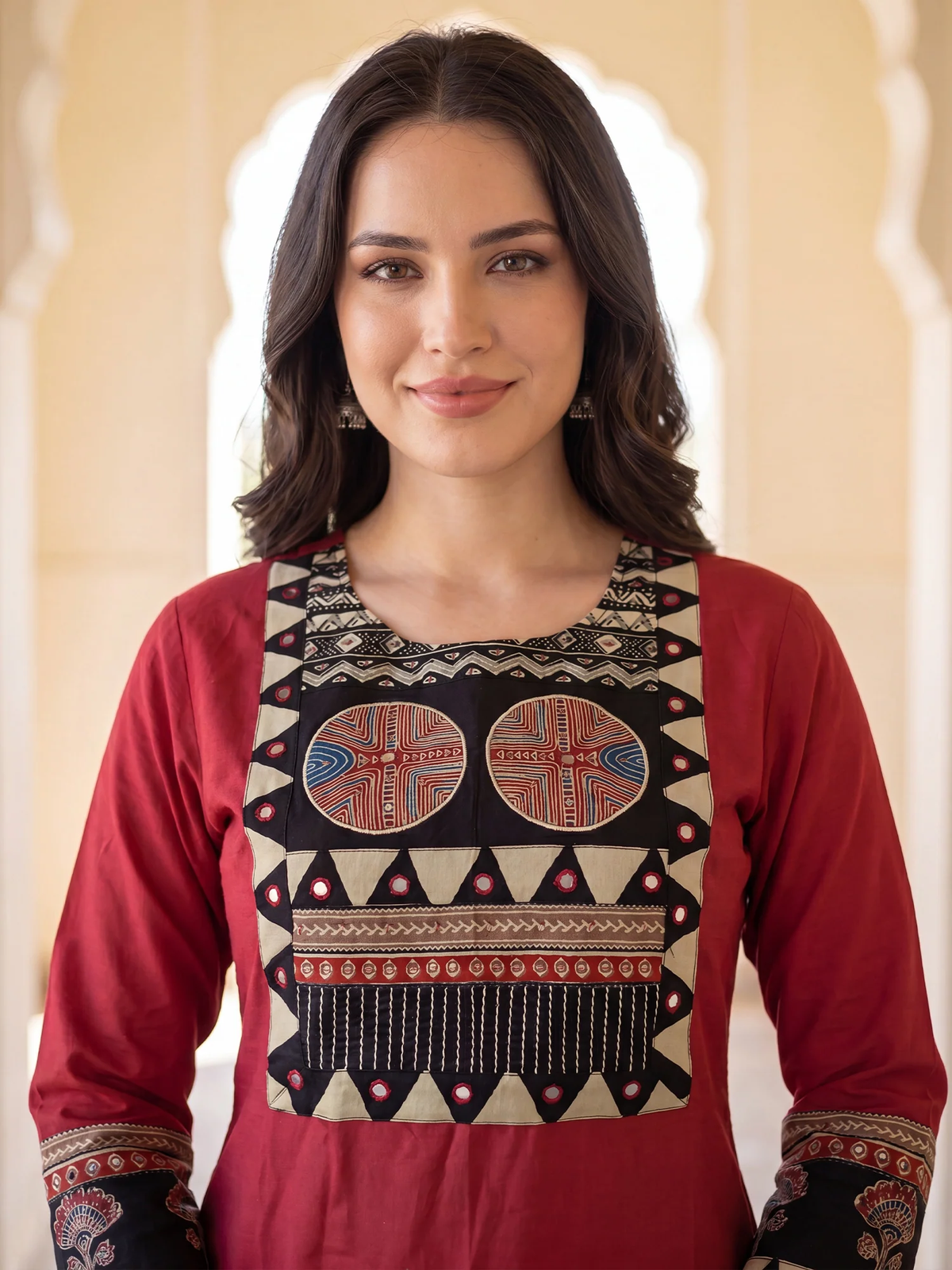A woman wearing a red long-sleeve kurta with a large beige embroidered yoke featuring geometric patterns and two circular motifs, set against a soft beige architectural background.