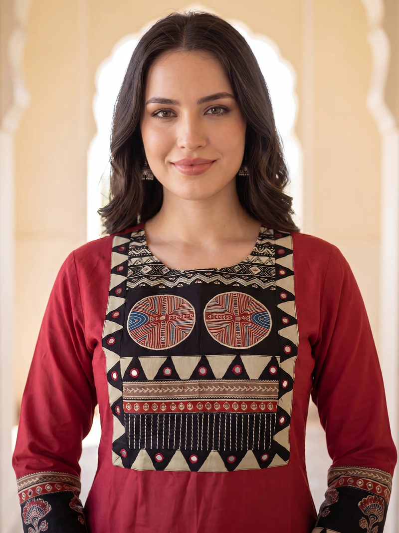 A woman wearing a red long-sleeve kurta with a large beige embroidered yoke featuring geometric patterns and two circular motifs, set against a soft beige architectural background.