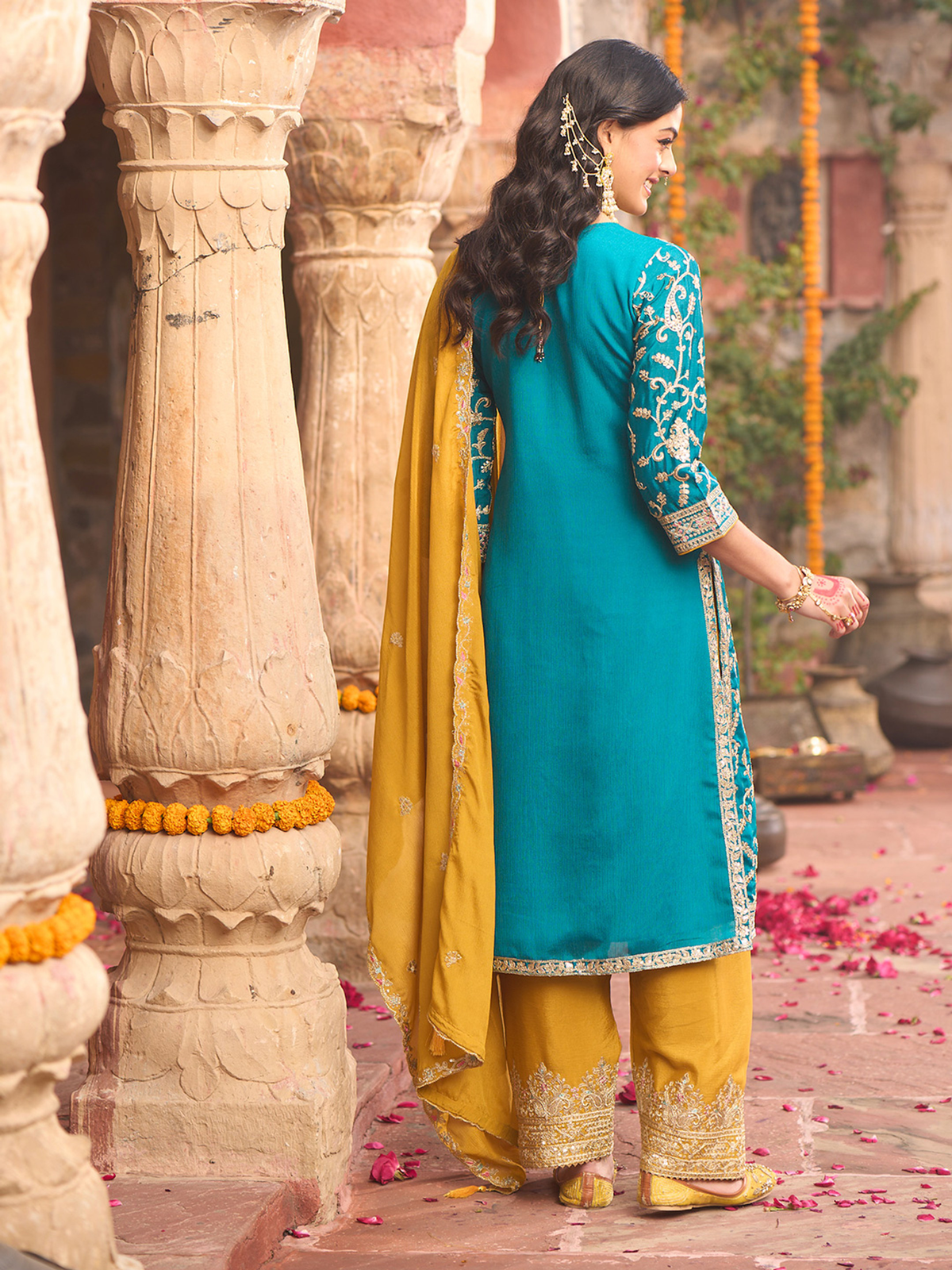 Woman in a teal and yellow traditional outfit standing in front of stone pillars with floral decorations.