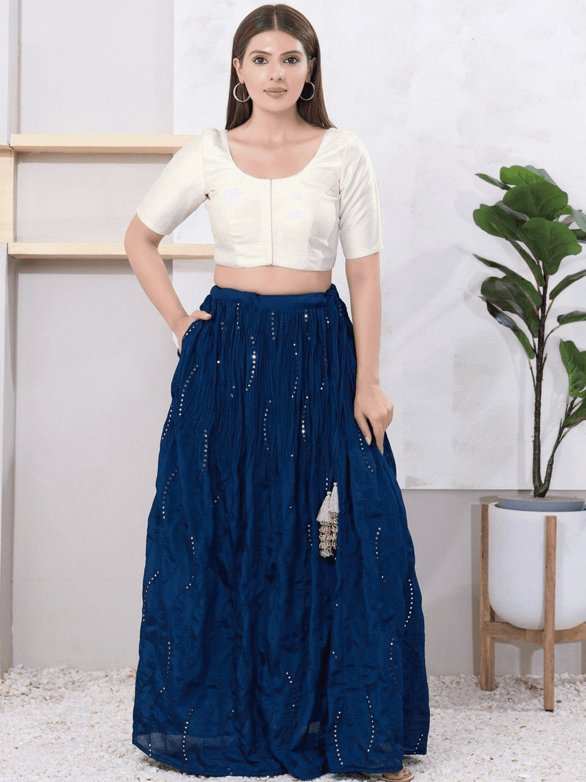 A woman modeling a navy blue maxi skirt with silver embellishments and a cream-colored short-sleeve crop top, standing in a styled indoor setting with a potted plant.