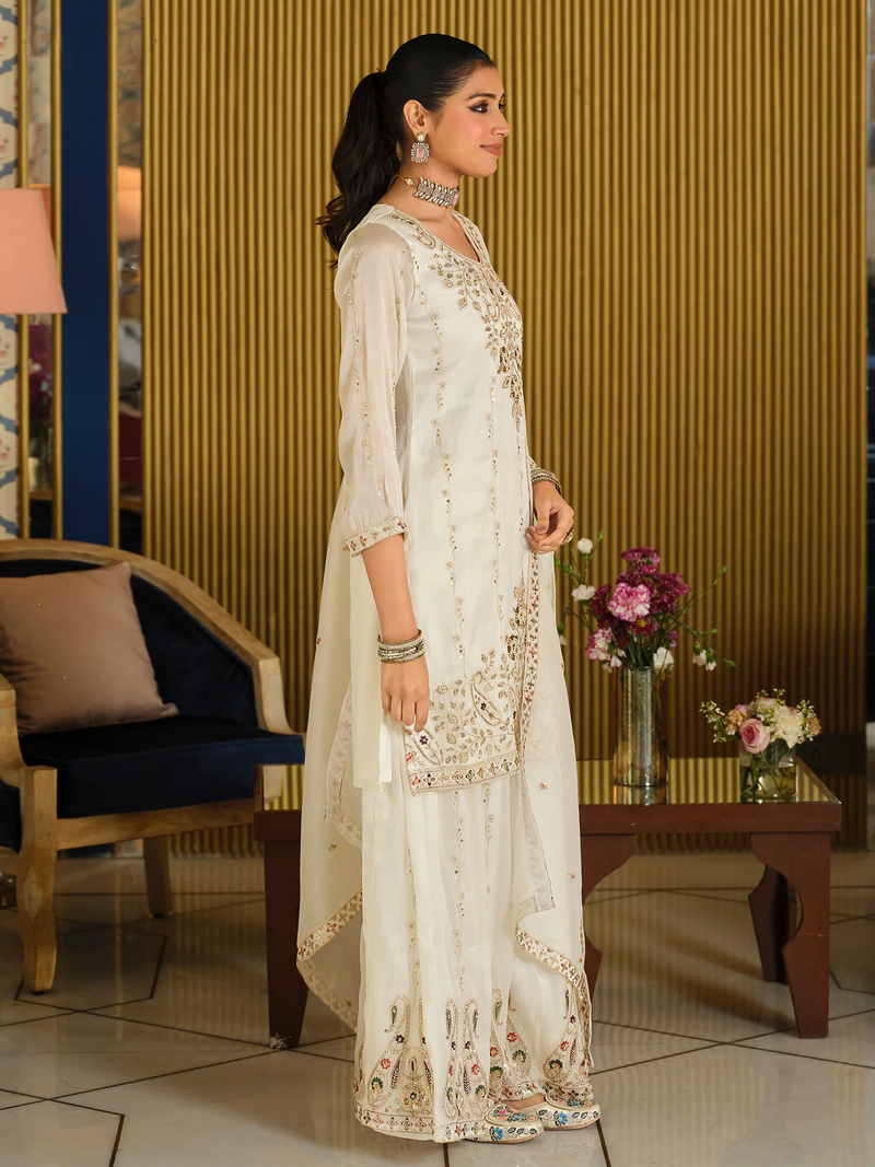 Woman in a white embroidered outfit standing in a decorated room with floral arrangements.