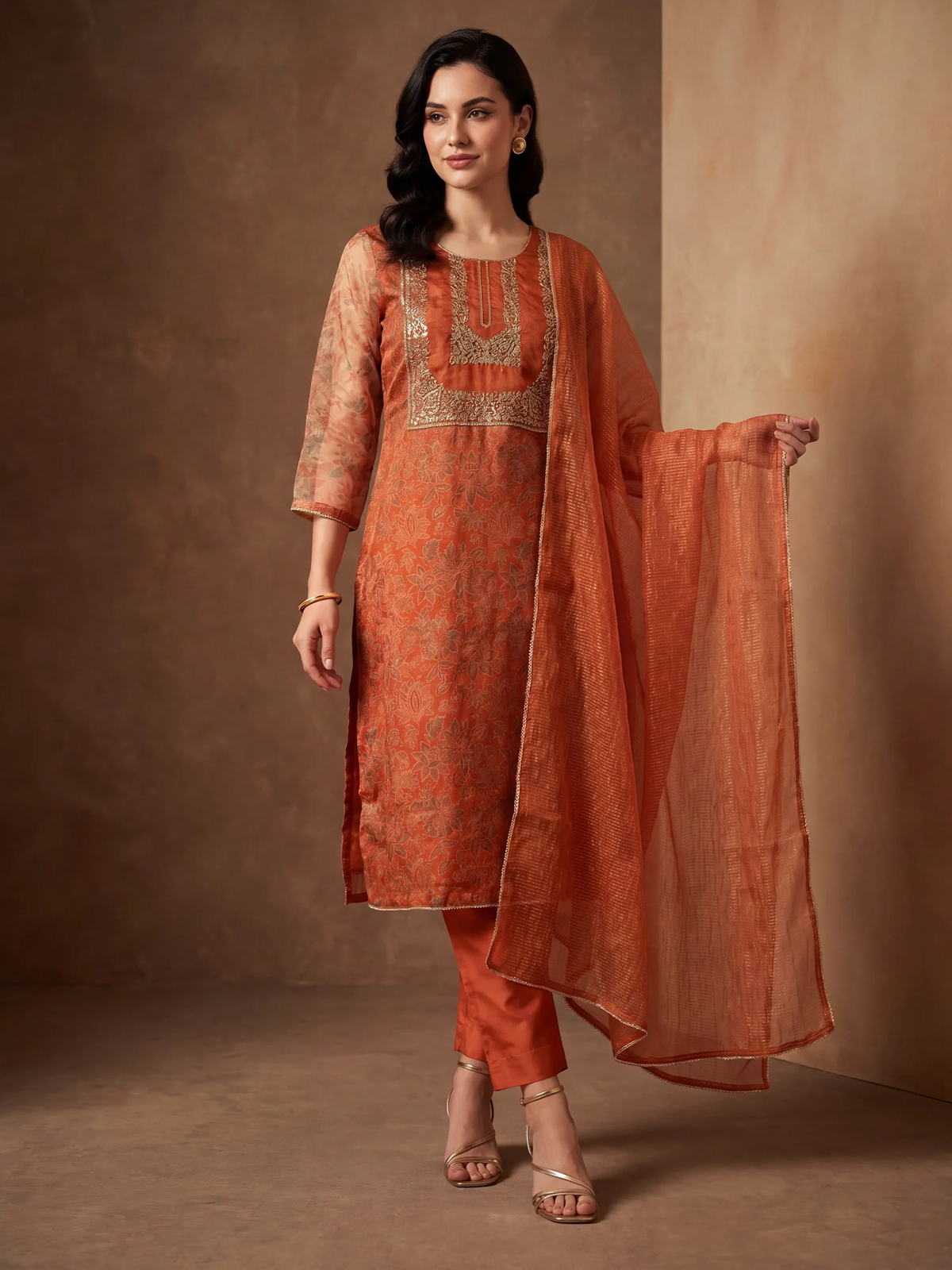 A woman models a traditional Indian outfit consisting of an orange kurta with gold embroidery, matching orange trousers, and a sheer orange dupatta, set against a warm brown studio backdrop.