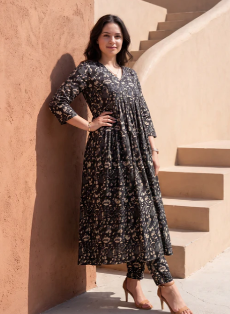 A woman wearing a black floral print maxi dress and matching pants stands outdoors against a warm beige stucco wall and steps.