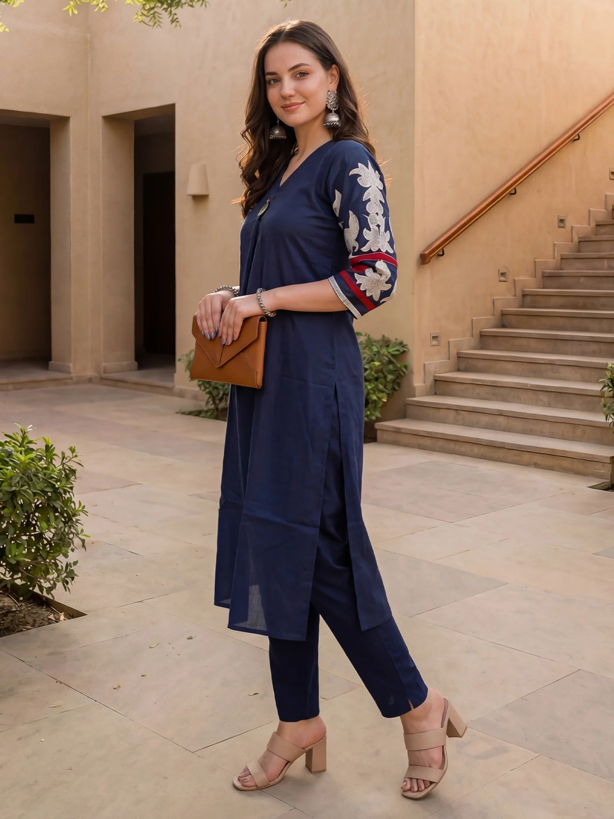 A woman models a navy blue three-piece kurta set with embroidered sleeves, holding a brown clutch bag in an outdoor courtyard setting.