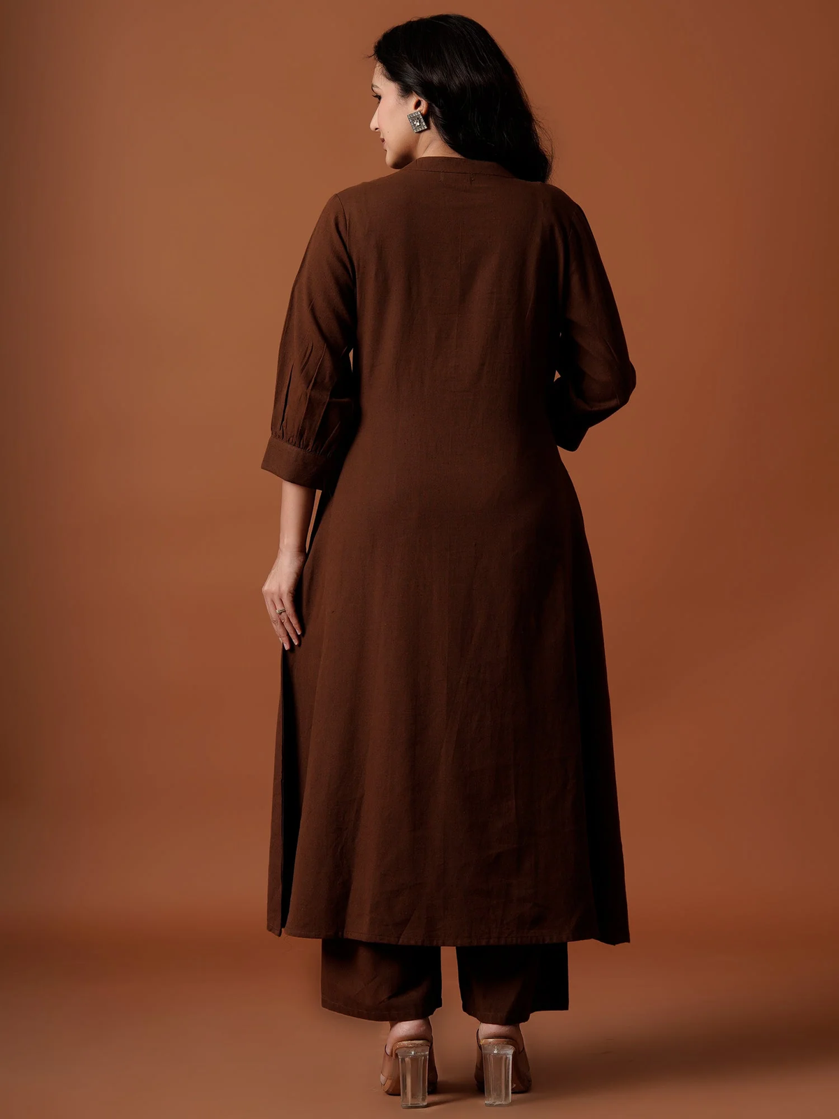A woman viewed from the back wearing a dark brown long kurta with three-quarter sleeves and matching brown pants, standing against a solid warm brown studio backdrop.