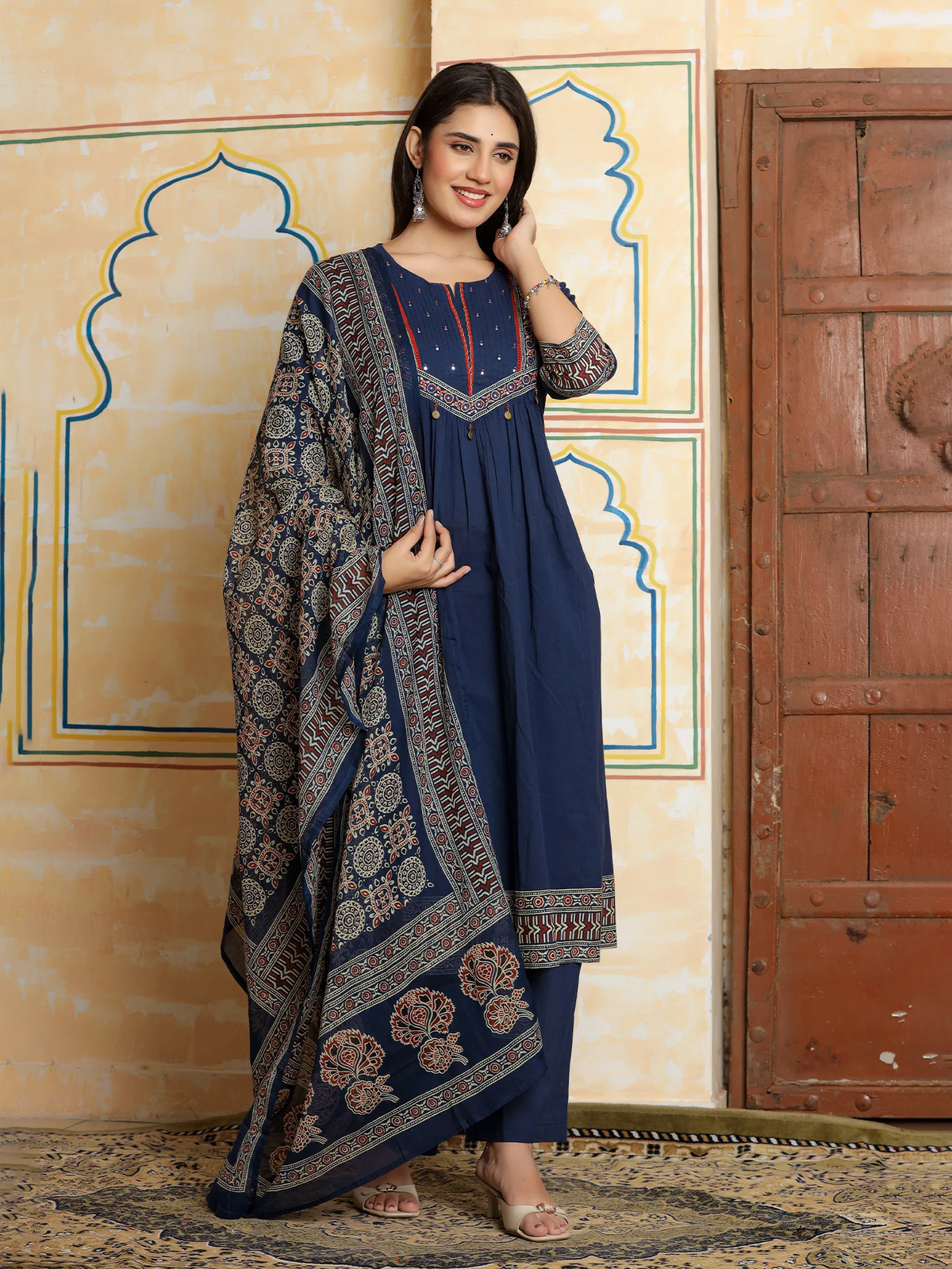 Woman in a blue dress with a patterned shawl standing in front of a decorative wall.