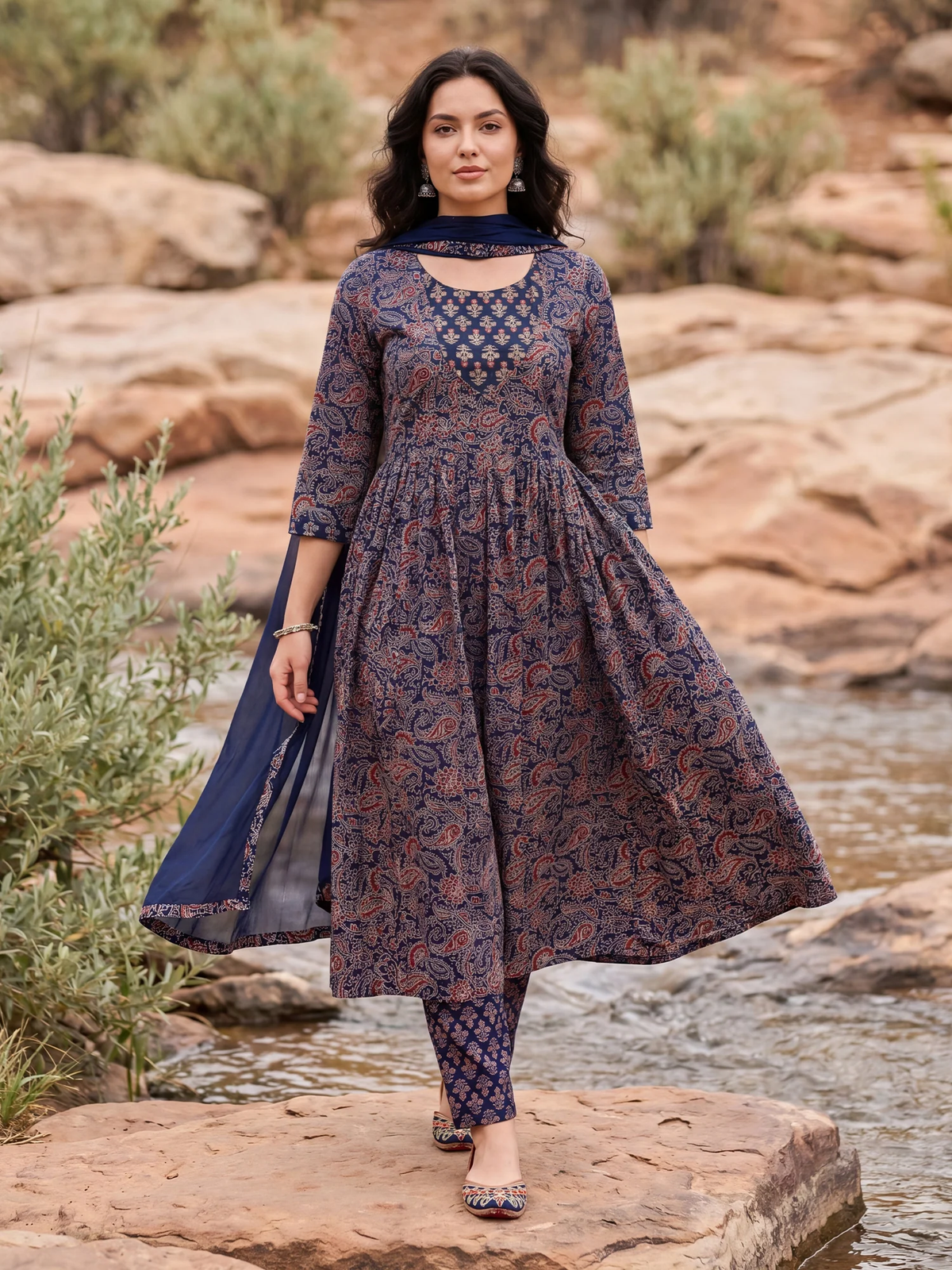 Woman in a blue and brown patterned outfit standing by a stream with rocks and greenery.