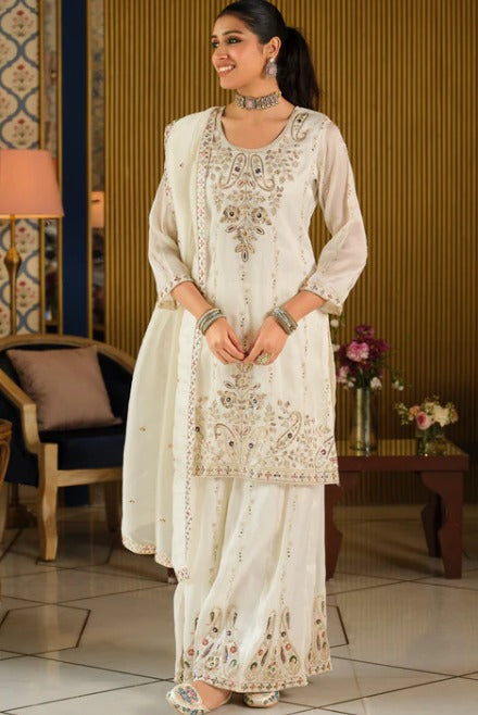 A woman models a traditional white three-piece salwar kameez suit with intricate gold and silver embroidery, set against an indoor background with a wooden slatted wall and decorative furniture.