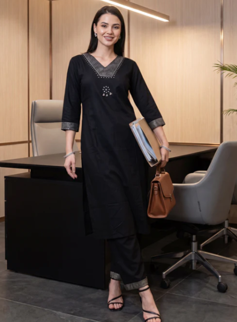 A woman modeling a black kurta set with gray embroidered details on the neckline and cuffs, standing in an office environment next to a desk and chair.