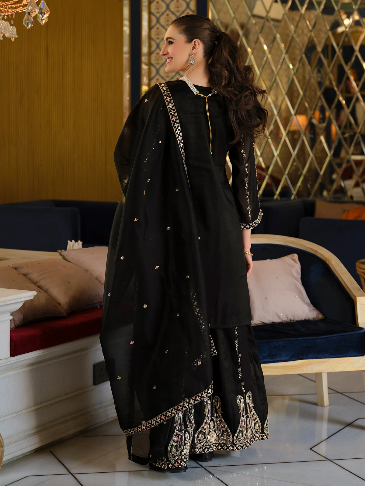 A woman modeling a black traditional South Asian outfit with a matching dupatta, featuring silver mirror work embroidery on the borders and sleeves, set in an elegant indoor lounge environment.