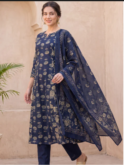 A woman models a navy blue three-piece traditional outfit with beige floral prints, consisting of a long kurta, trousers, and a matching dupatta, set against a beige stucco wall background.