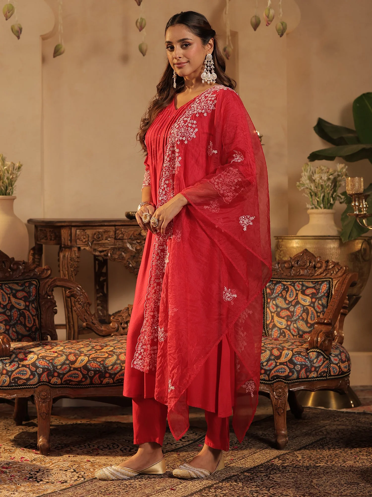 A woman modeling a red traditional Indian salwar kameez suit with white floral embroidery, standing in an ornate room with patterned sofas and decorative plants.