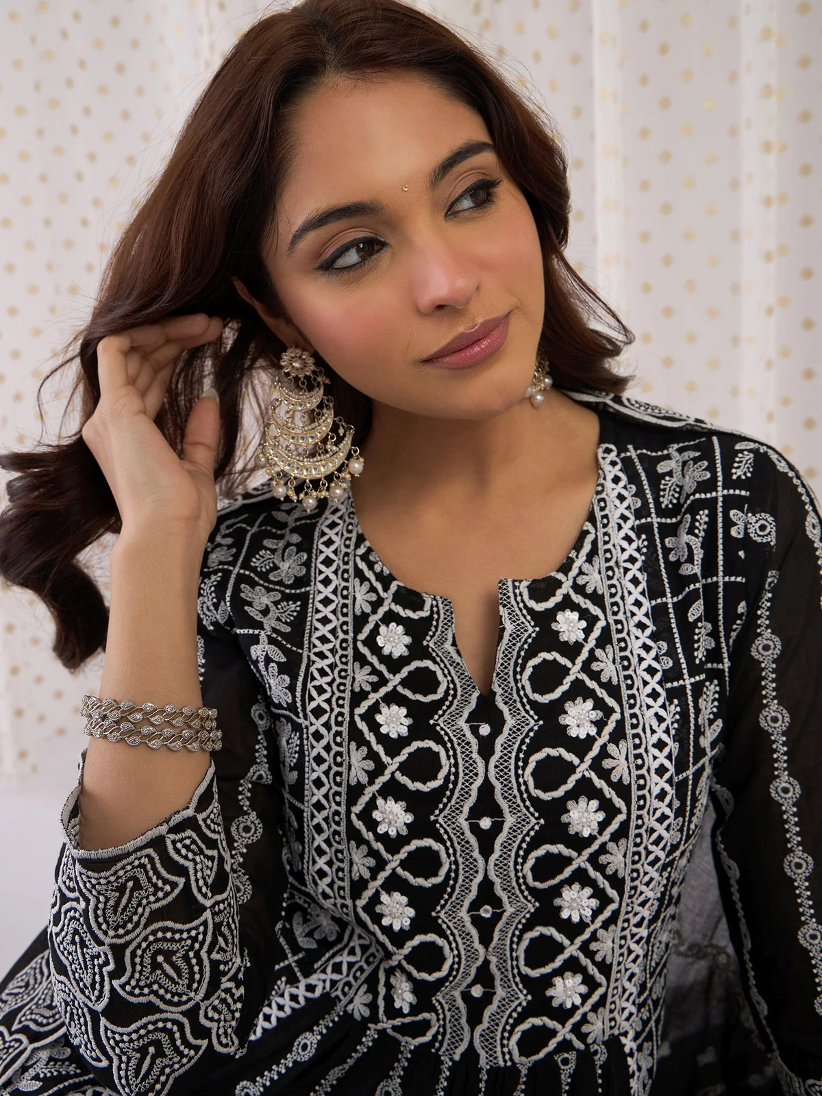 Woman wearing a black and white embroidered traditional outfit with jewelry against a light background.