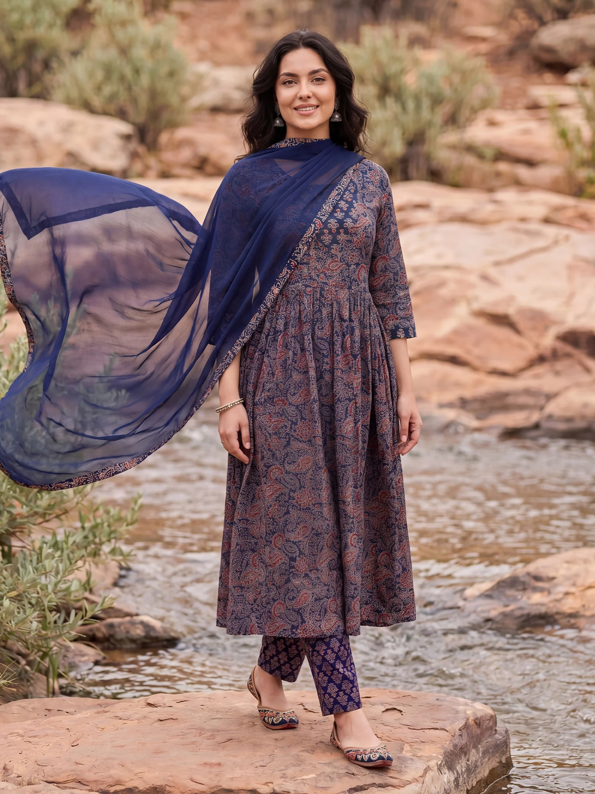A woman stands outdoors by a rocky stream wearing a long-sleeved printed kurta in a muted purple and brown paisley pattern, paired with matching patterned pants and a flowing navy dupatta.