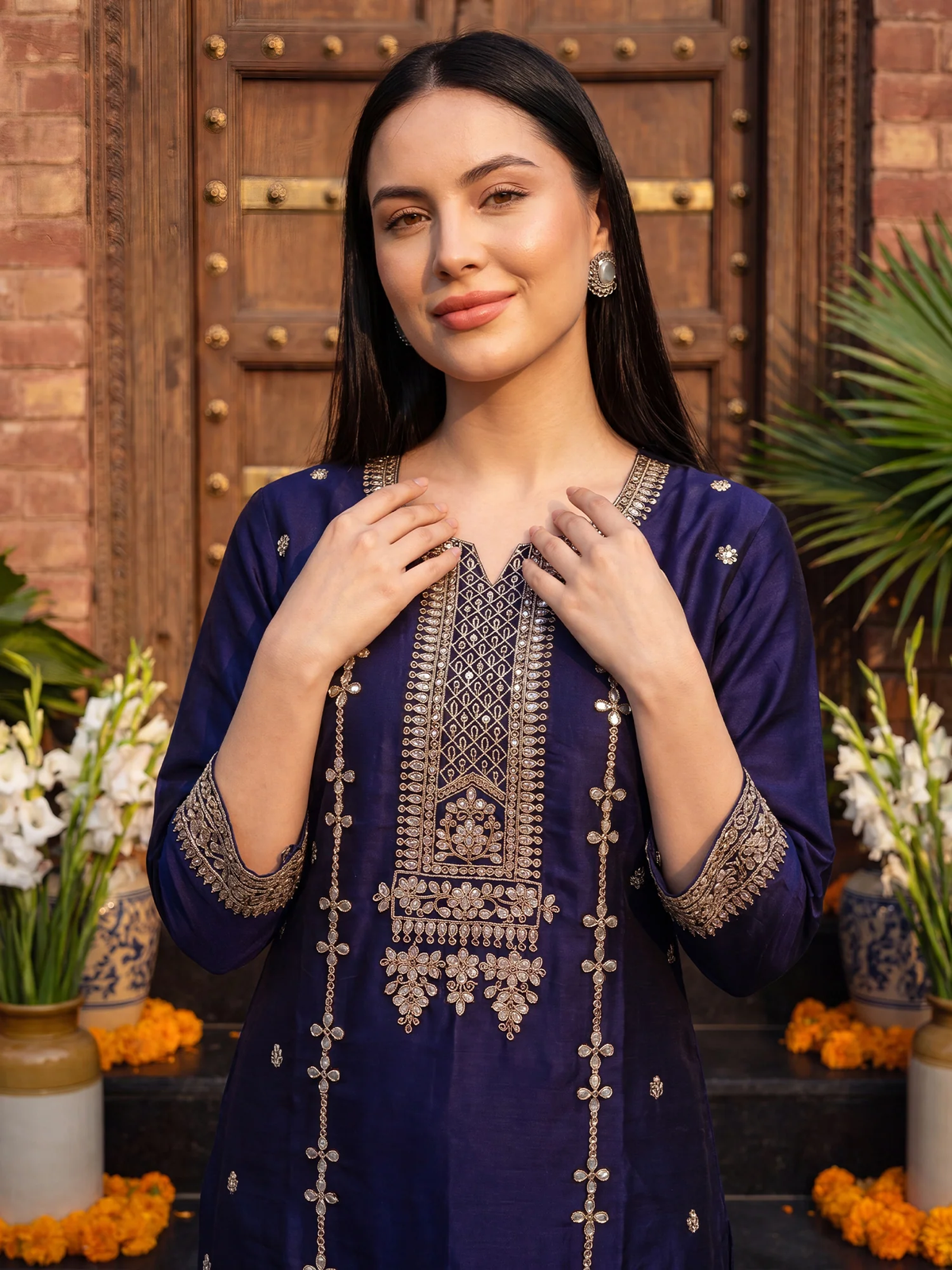 A woman wearing a navy blue embroidered kurta with intricate gold and silver metallic detailing on the neckline, sleeves, and front drape, standing in front of a rustic wooden door with decorative plants and flowers.