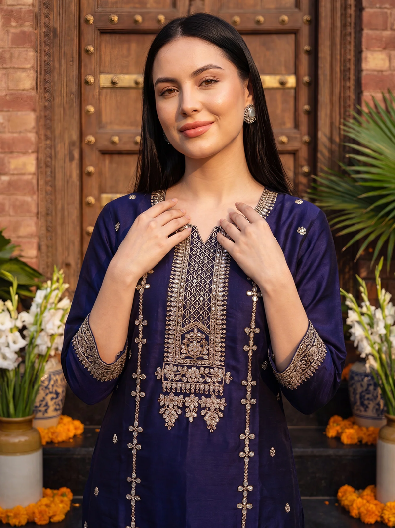 A woman wearing a navy blue embroidered kurta with intricate gold and silver metallic detailing on the neckline, sleeves, and front drape, standing in front of a rustic wooden door with decorative plants and flowers.