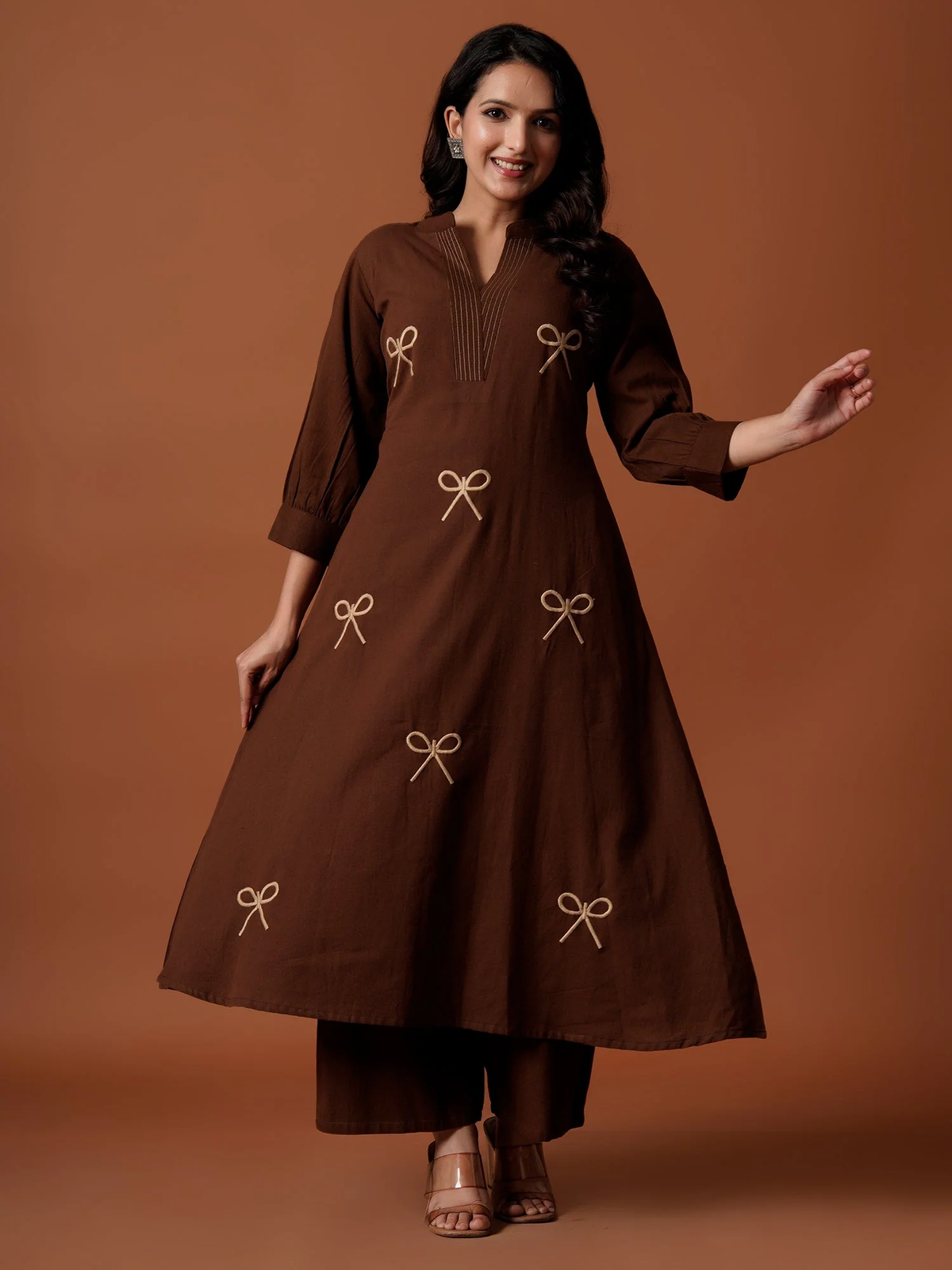 A woman modeling a brown A-line kurta with beige embroidered bow motifs, paired with matching brown trousers, set against a solid warm brown studio background.