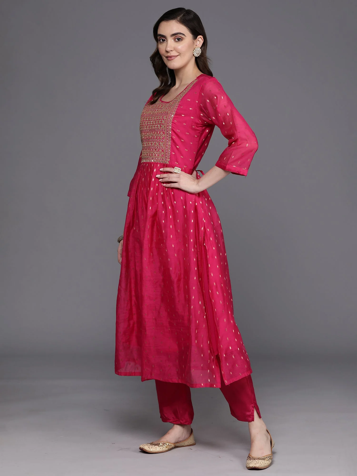 A woman modeling a bright pink traditional Indian outfit consisting of a kurta with a beige embroidered yoke, matching pants, and flat beige shoes, set against a solid gray studio background.