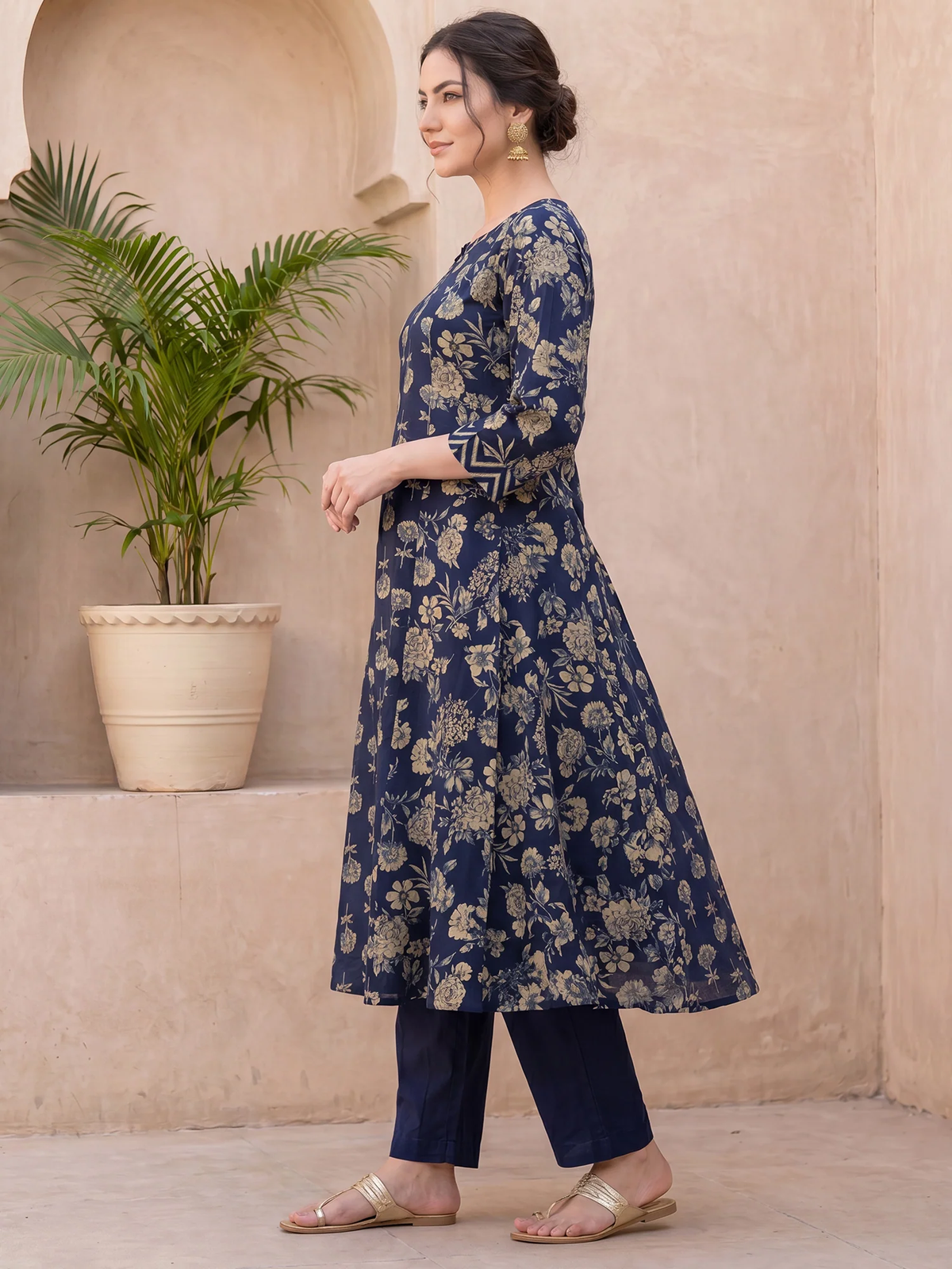Woman wearing a blue floral dress standing in front of a beige wall with a plant.