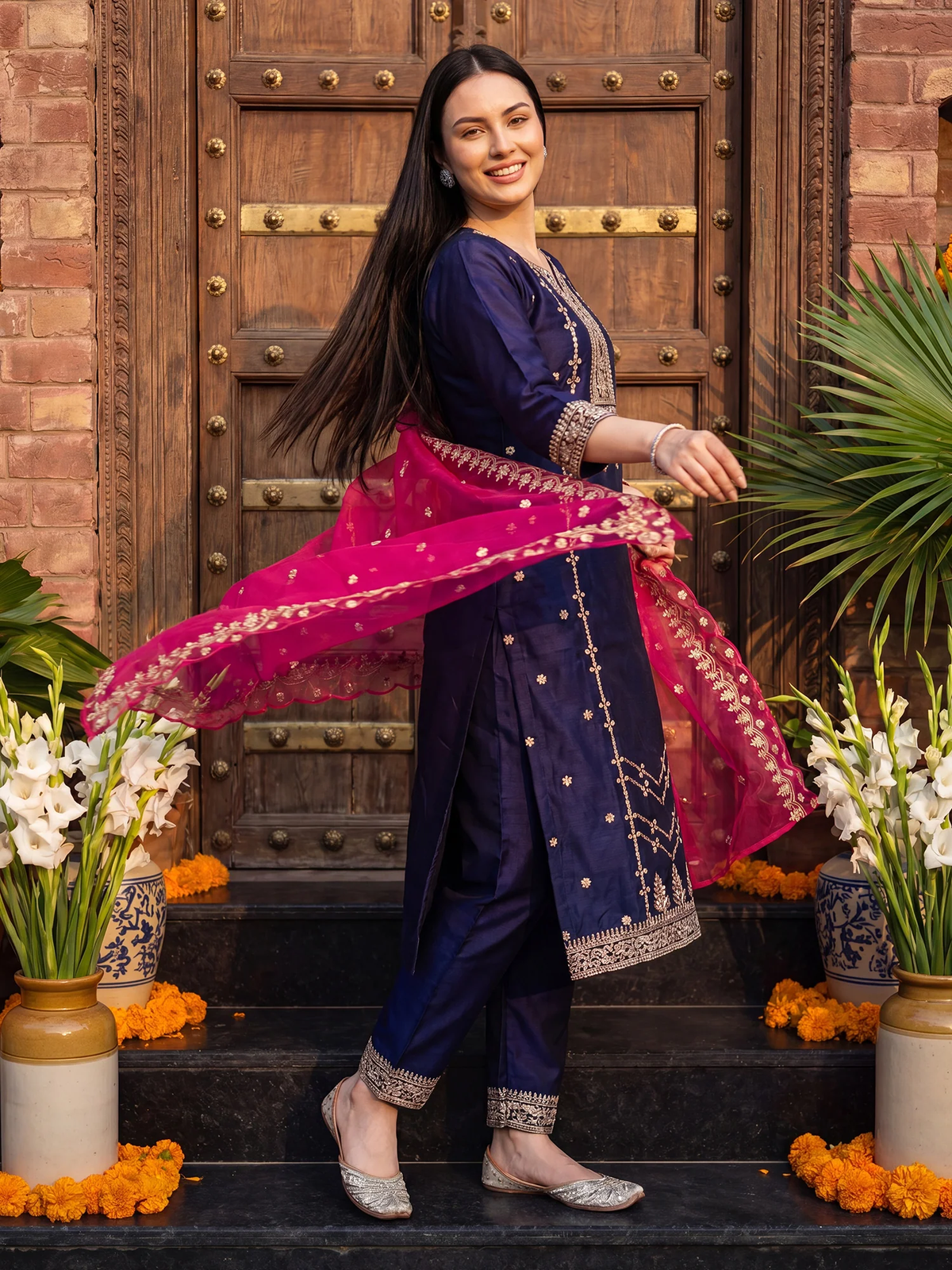 A woman modeling a traditional Indian outfit consisting of a navy blue kurta and trousers with gold embroidery, paired with a bright pink dupatta, standing in front of a rustic wooden door.