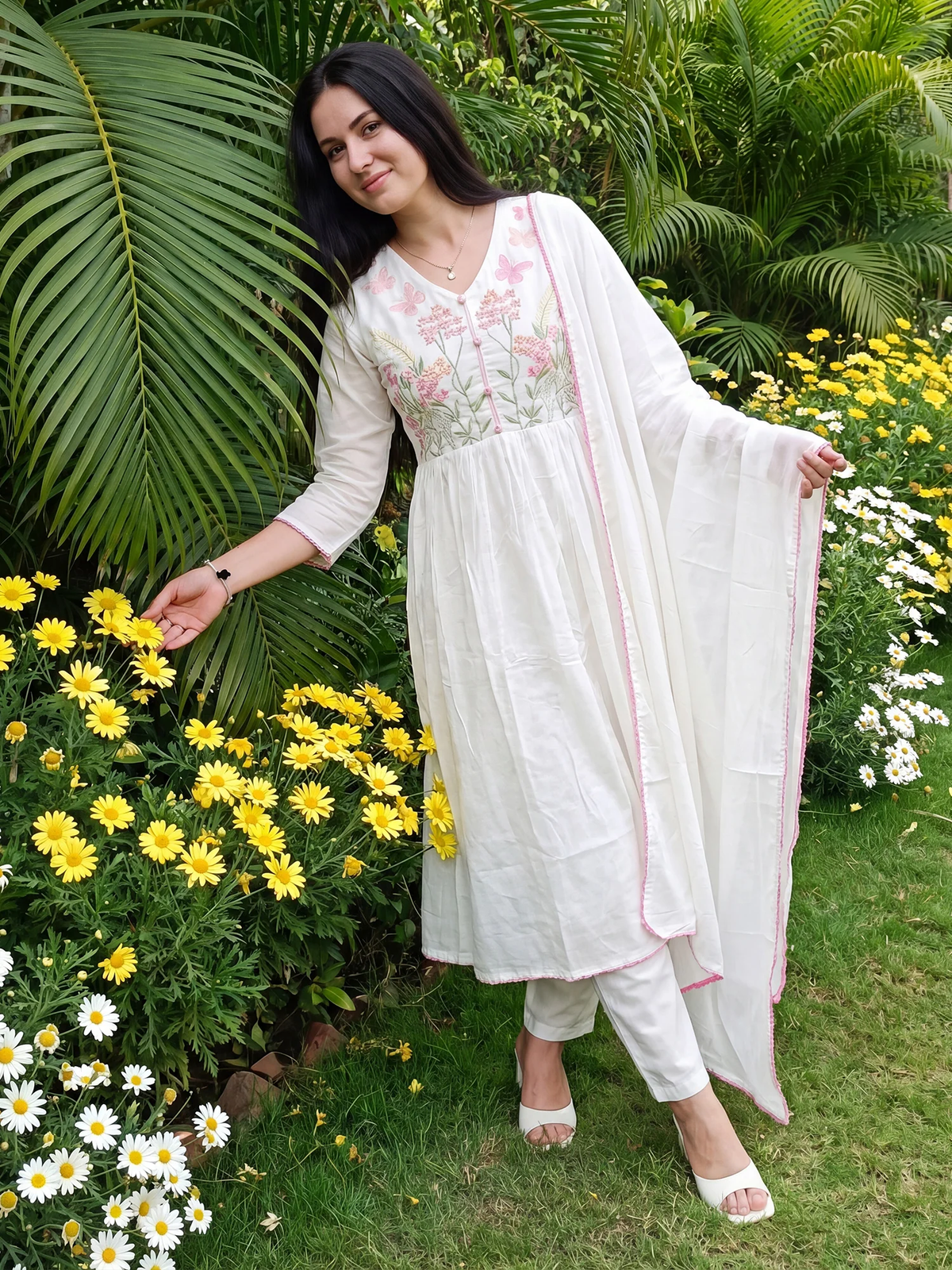 Woman in a white traditional outfit standing among flowers and greenery