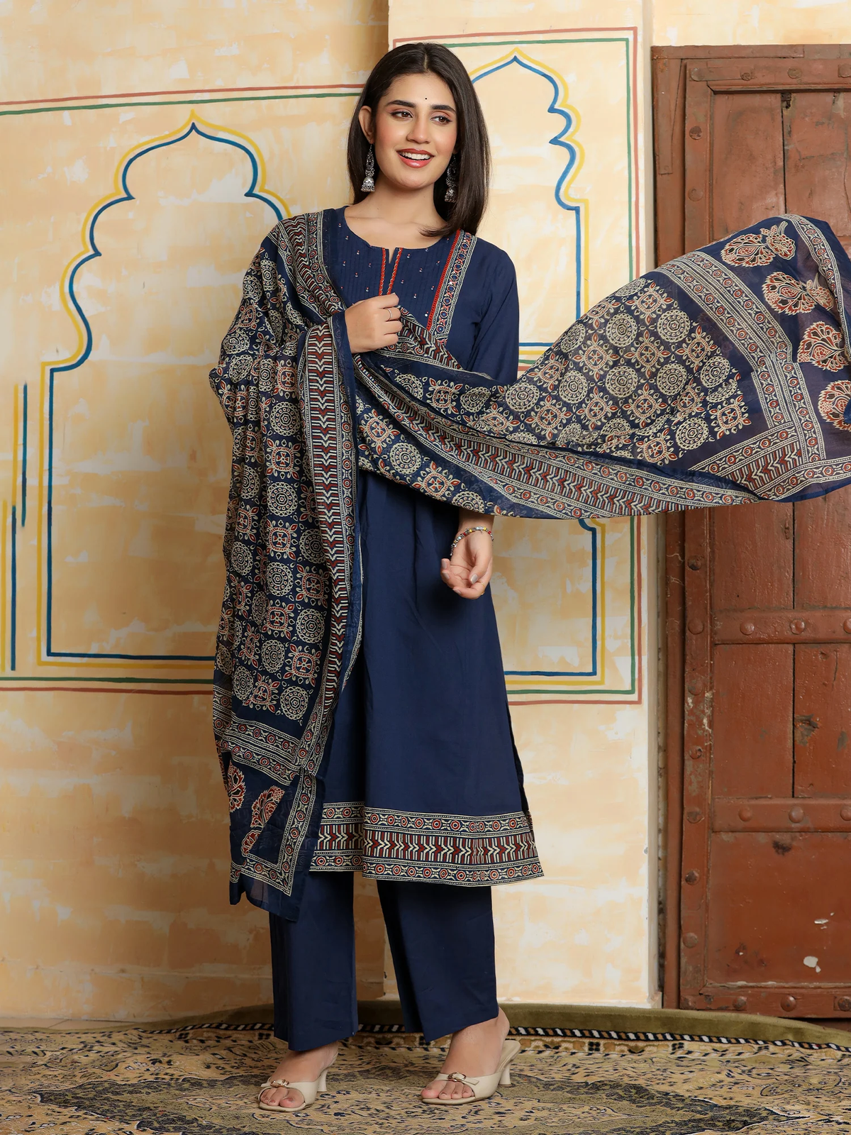 A woman models a navy blue three-piece salwar kameez suit with matching printed dupatta in front of a beige wall with traditional arch designs.