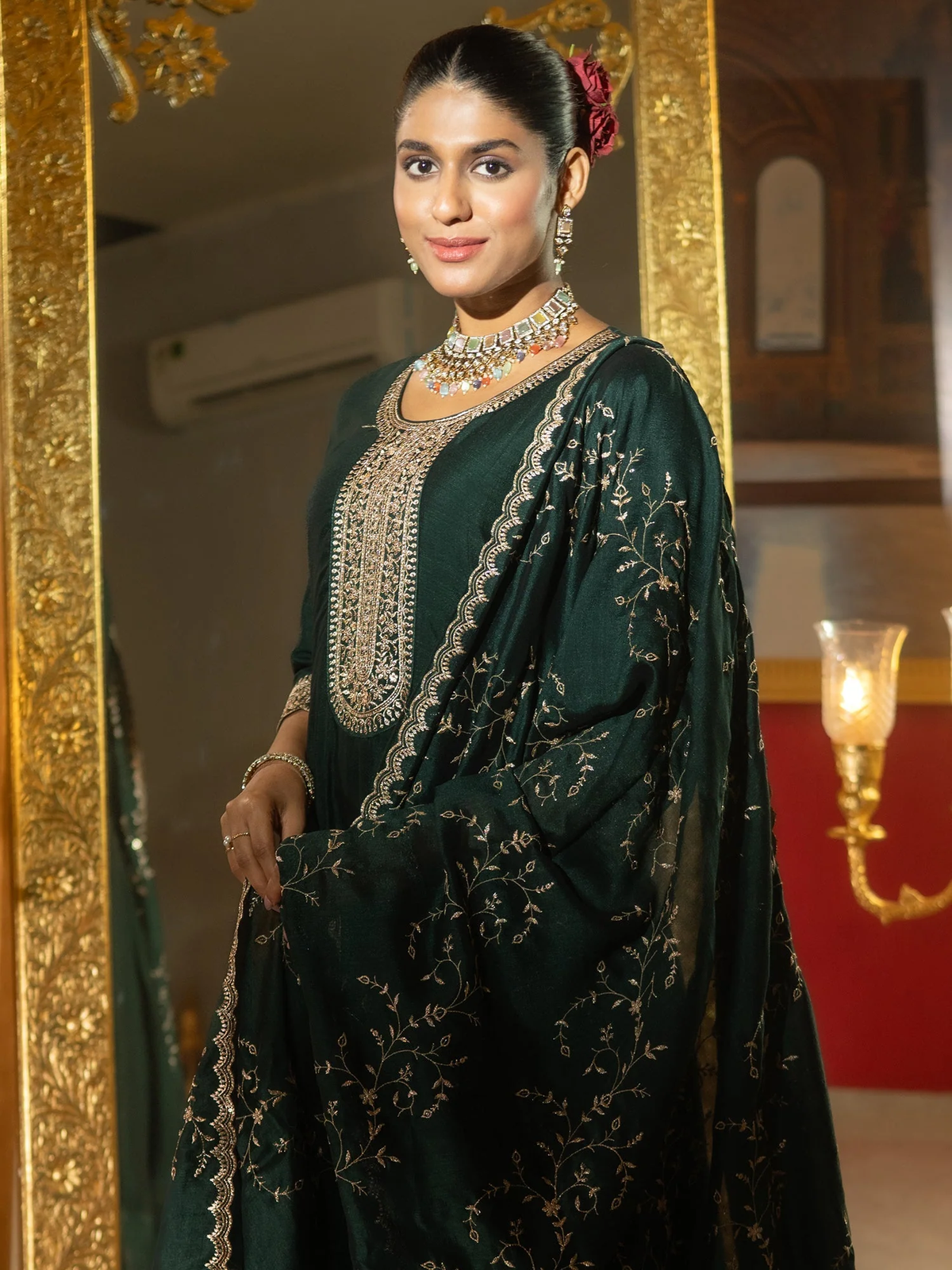 Woman in a dark green traditional outfit with intricate patterns, standing indoors.