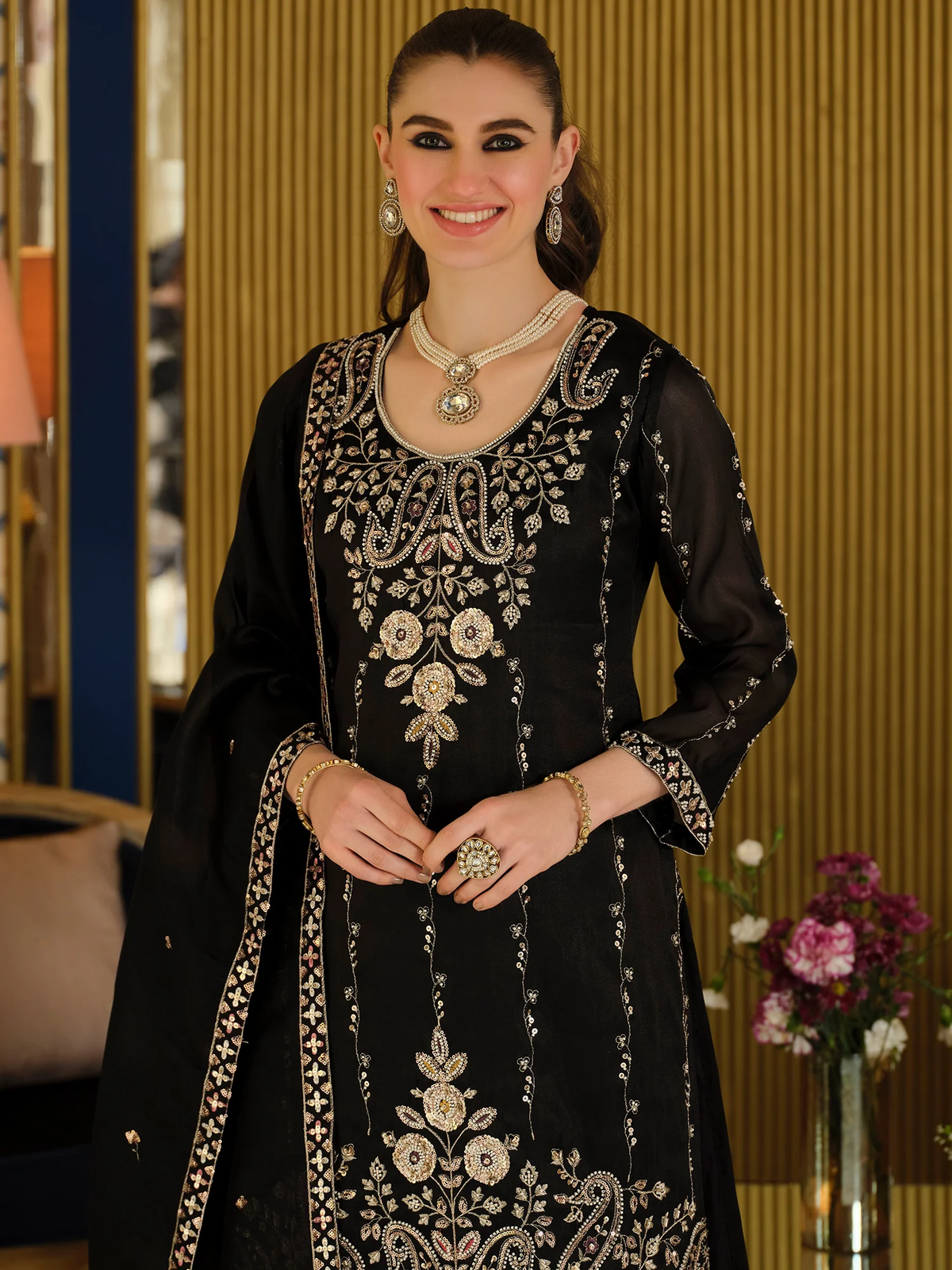 A woman wearing a black traditional embroidered outfit with ornate gold and silver floral and paisley detailing, standing in an indoor setting with a warm wood-paneled background.