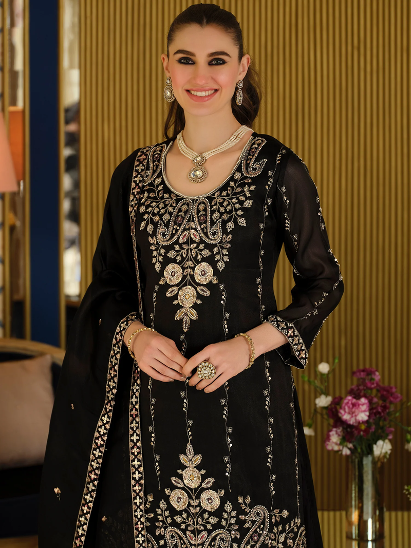 A woman wearing a black traditional embroidered outfit with ornate gold and silver floral and paisley detailing, standing in an indoor setting with a warm wood-paneled background.