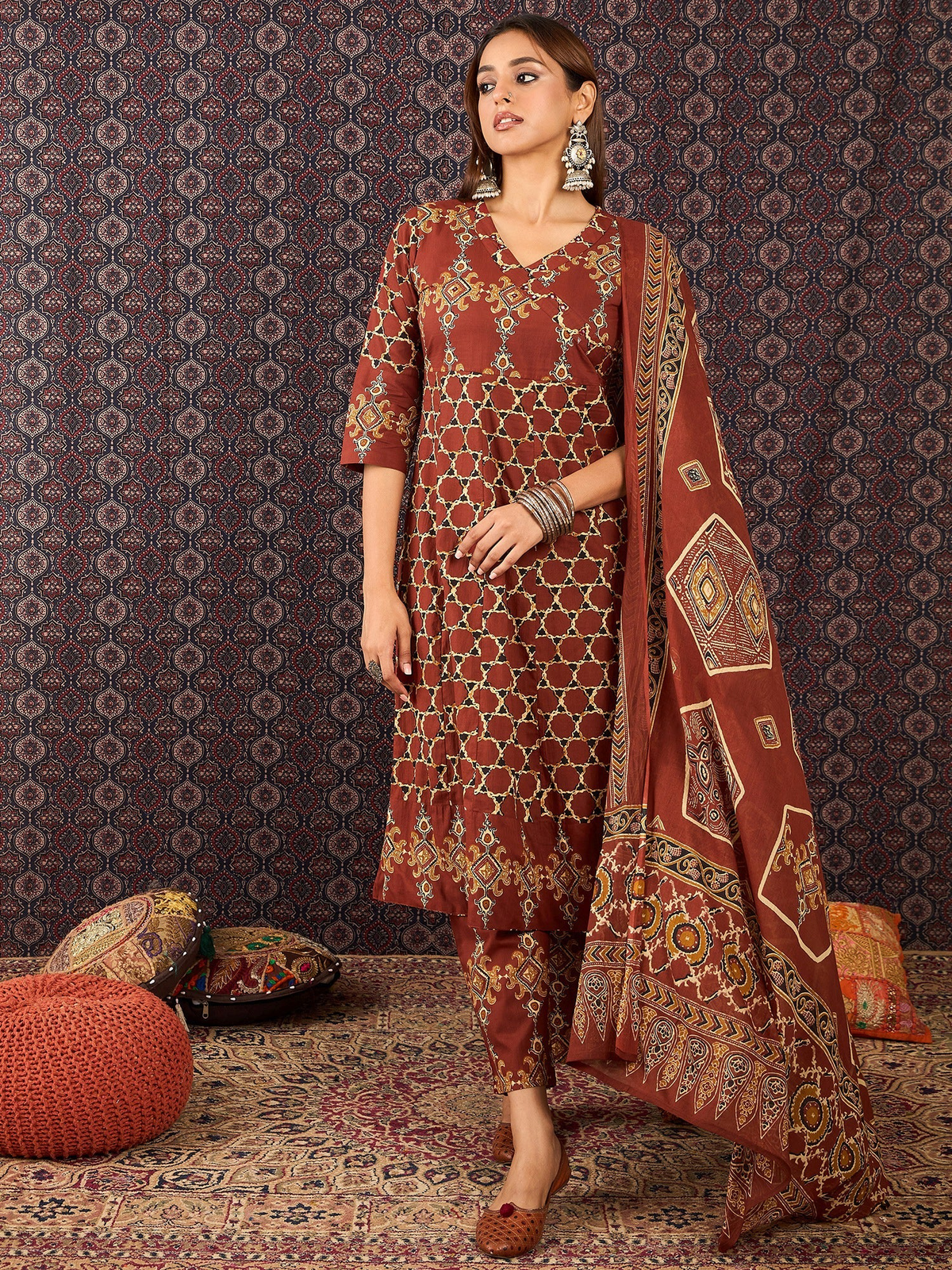 Woman wearing a traditional maroon and gold outfit with a matching dupatta against a patterned wall.