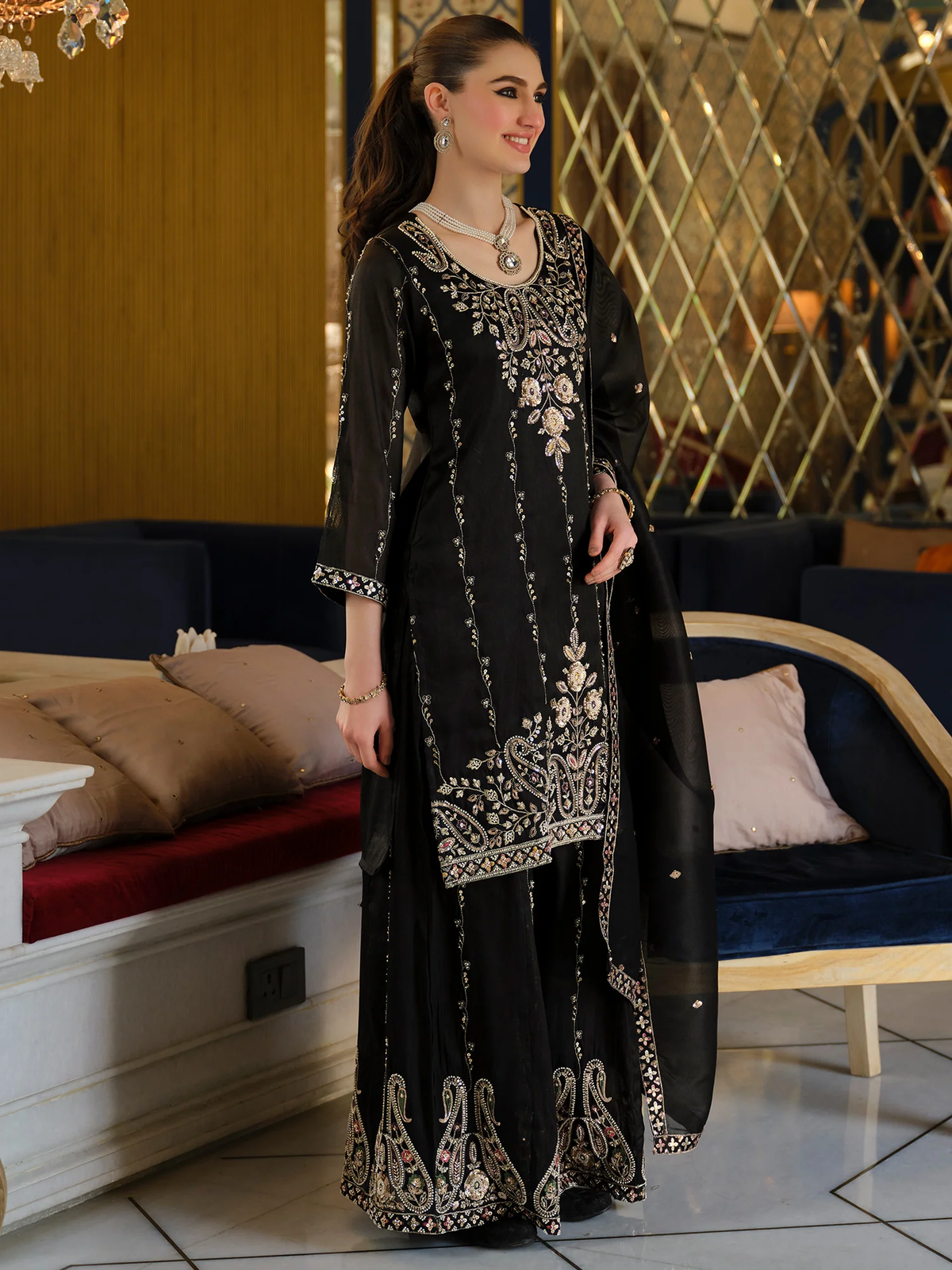 A woman models a black three-piece traditional outfit with intricate silver and gold embroidery, featuring a long kurta, matching flared pants, and a sheer dupatta, set against an indoor background with a sofa and decorative wall.