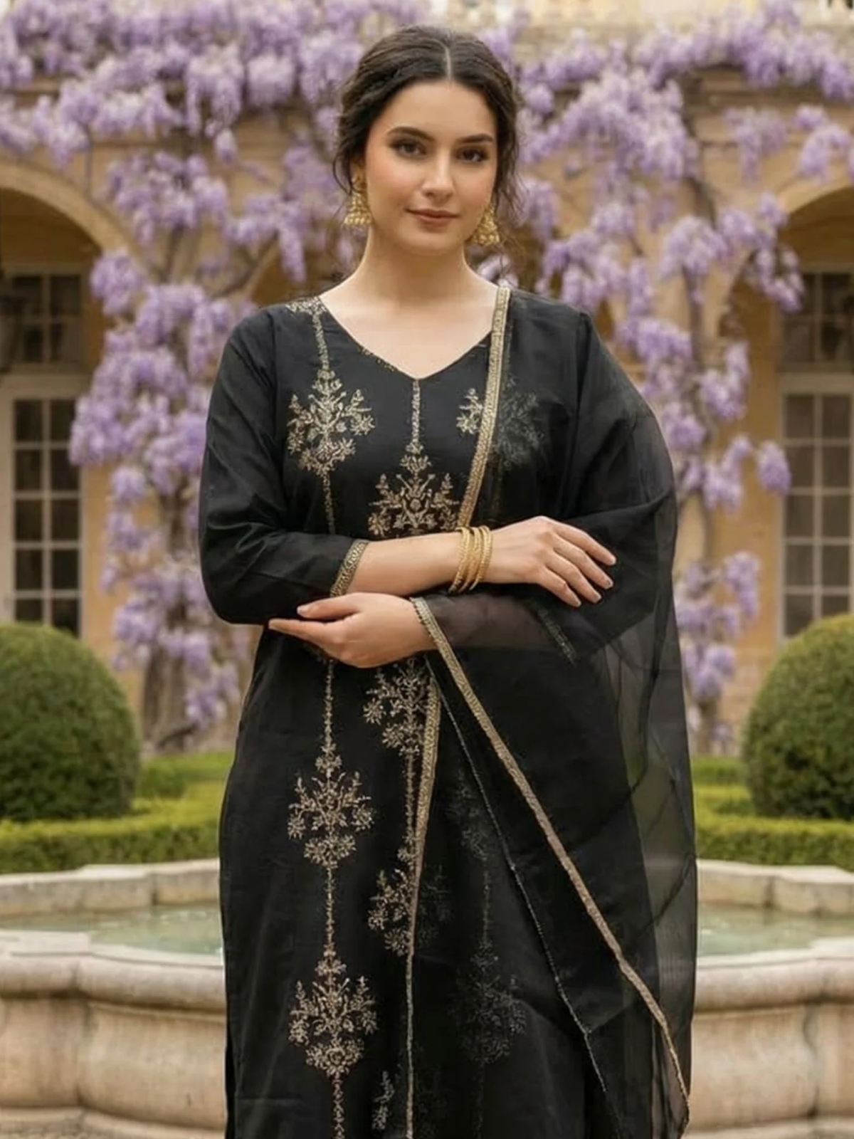 Woman in a black embroidered outfit standing in front of a building with purple flowers.