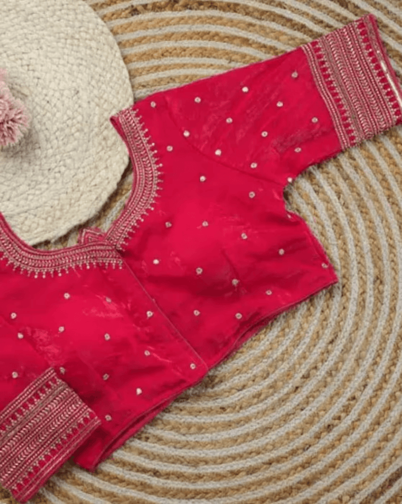 A bright pink traditional blouse with silver mirror work embellishments and gold border embroidery, laid flat on a woven straw mat next to a cream-colored woven hat.