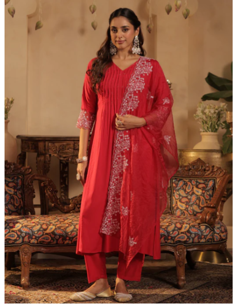 A woman modeling a red ethnic three-piece salwar kameez suit with white floral embroidery, standing in a decorated room with ornate wooden furniture.